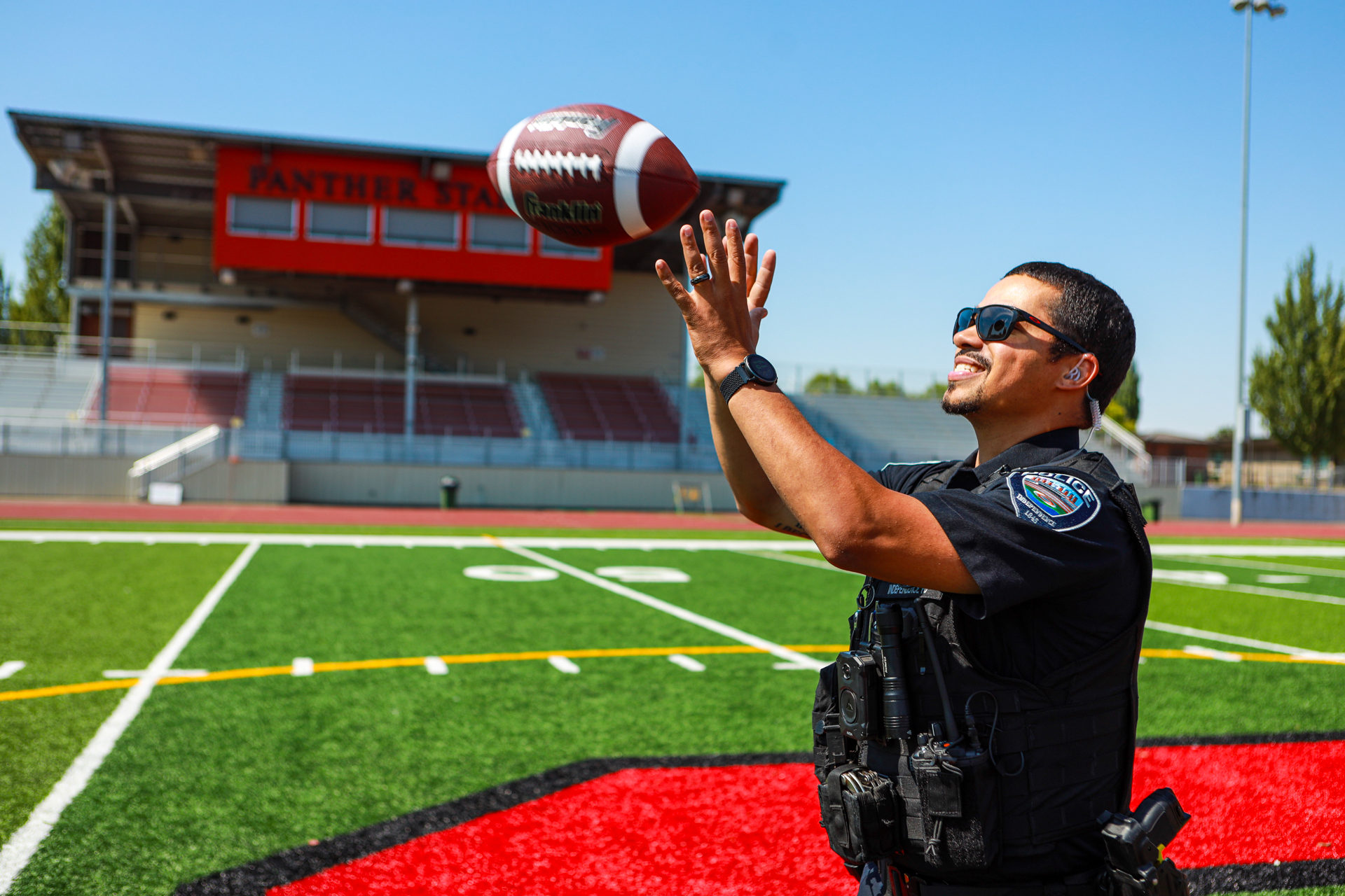officer preston playing with a football on a football field and smiling. the football stadium is behind him