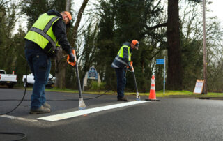 two public works crewmembers use thermal flame devices to heat thermoplastic lines onto the pavement.