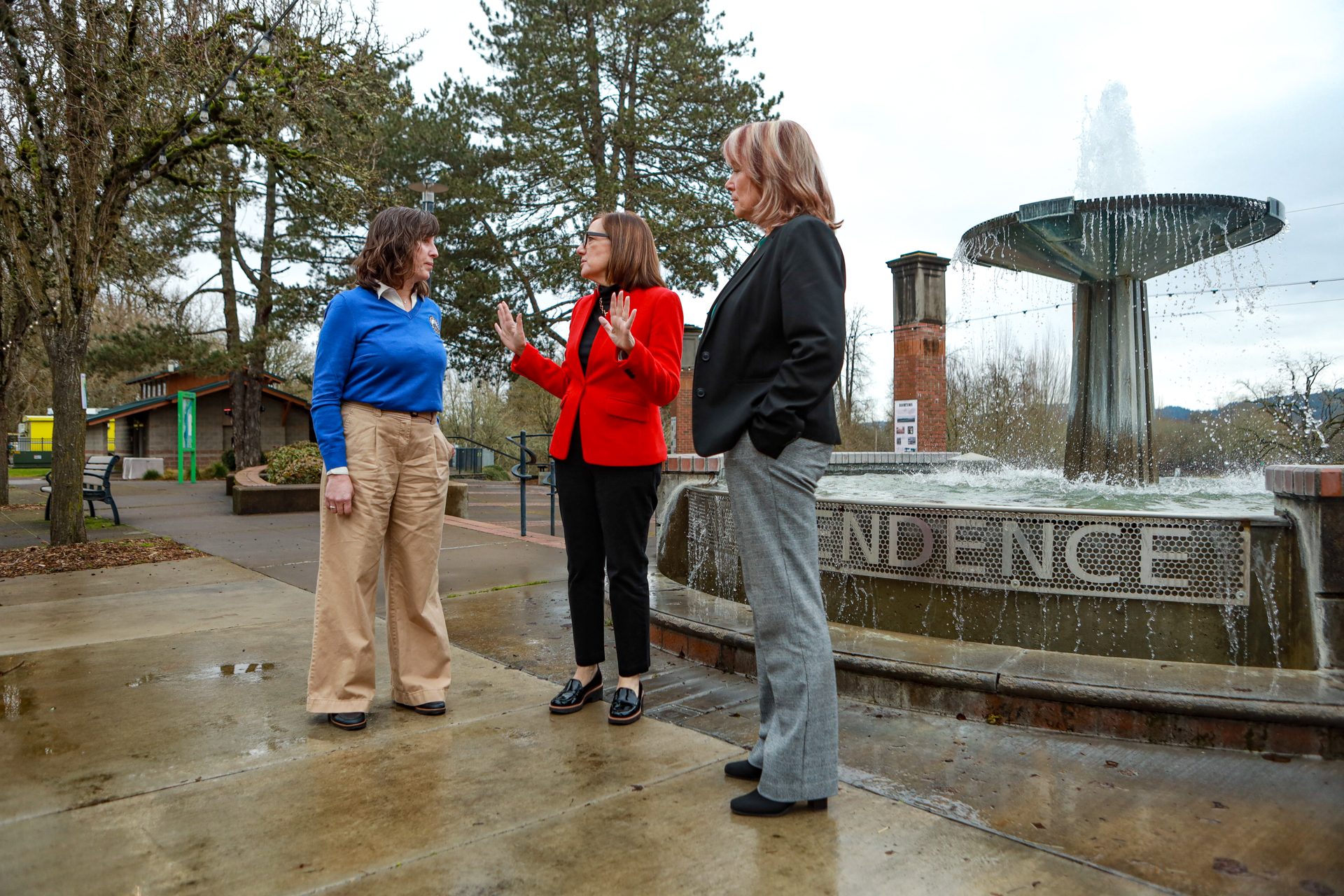 three women stand by a fountain reading "Independence" and engage in conversation. the woman in the middle, wearing a red blazer gestures with her hands as she talks.