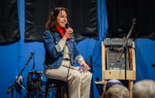 Mayor Kate sits on a stool holding a microphone, smiling as she speaks to attendees during a Town Hall meeting.