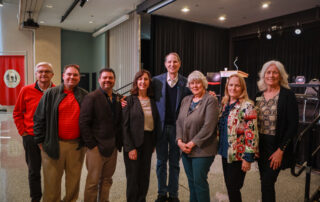 A group of eight people stand together in a well-lit auditorium, smiling warmly. The setting includes a stage with a podium and chairs.