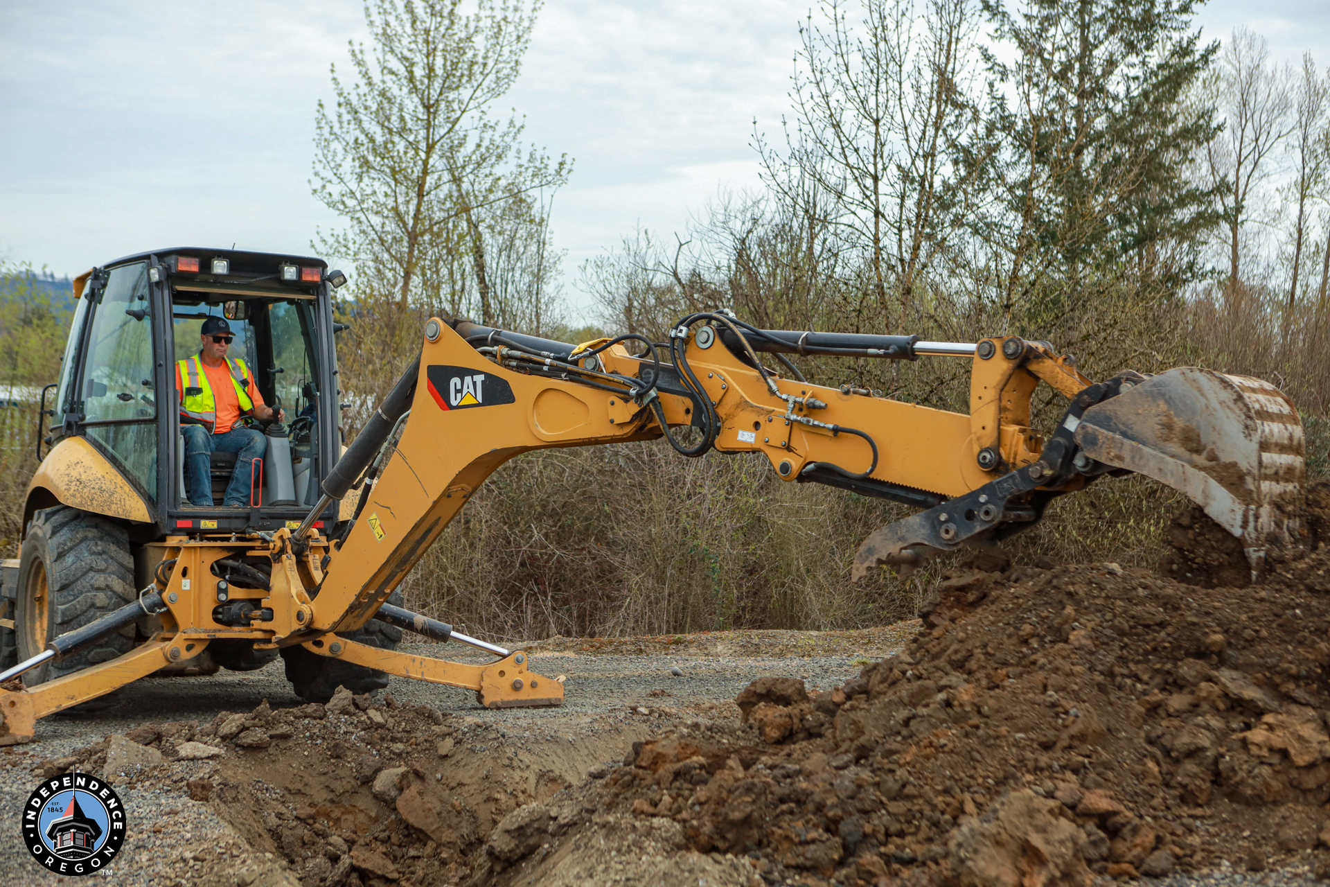 A construction worker in a safety vest operates a yellow backhoe, digging into a dirt mound on a cloudy day, with trees in the background.