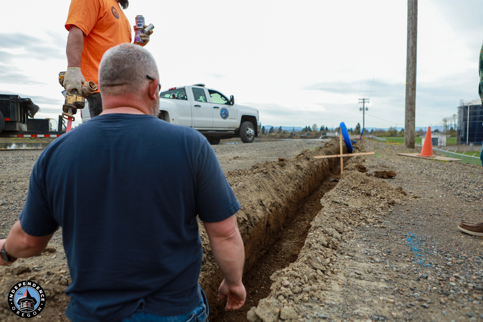 Two men work near a trench on a gravel road. One holds a tool, the other observes. A white truck and orange cone are in the background.