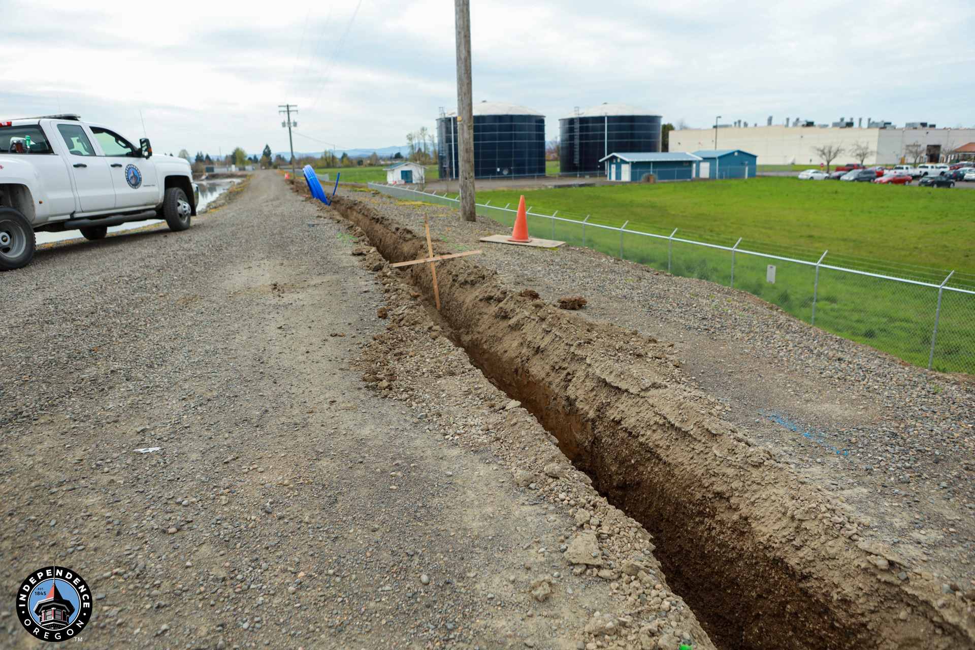 A gravel road with a long trench beside it. A white utility truck is parked nearby. Orange cones and a blue pipe are visible. Industrial tanks are in the background.