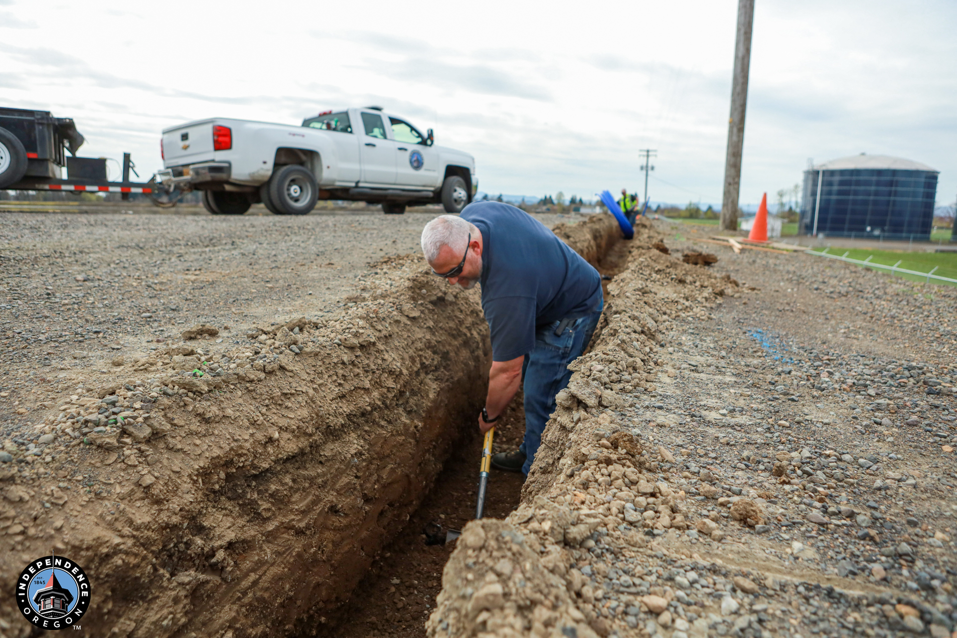 A man in work attire digs a trench on a gravel road near parked utility trucks. The scene conveys a focused, industrious atmosphere.