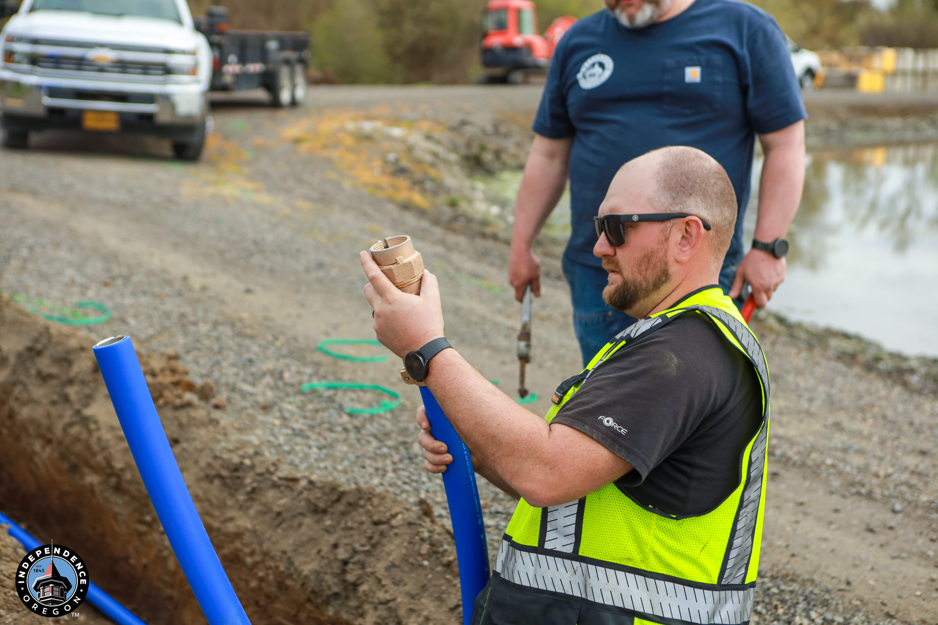 A construction worker in a safety vest examines a pipe fitting at a gravel worksite. Another worker stands nearby. Trucks are visible in the background.