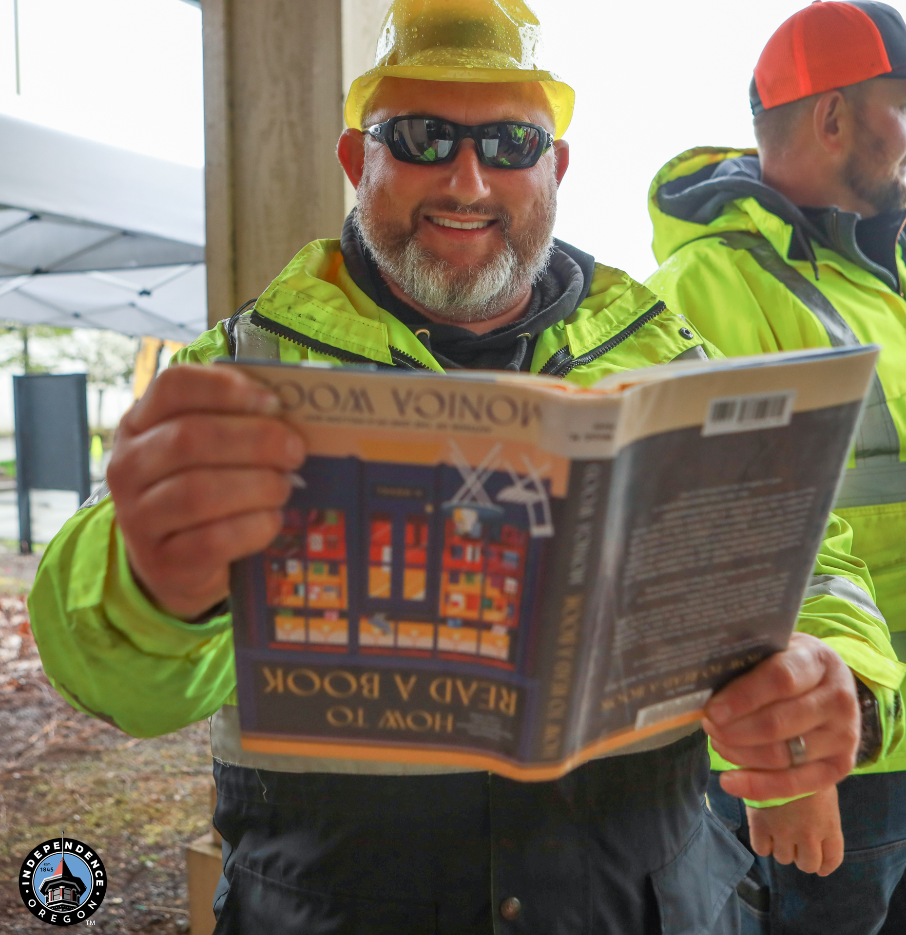 A man in a yellow hard hat and bright jacket smiles while holding a book upside down, exuding humor and lightheartedness at a construction site.
