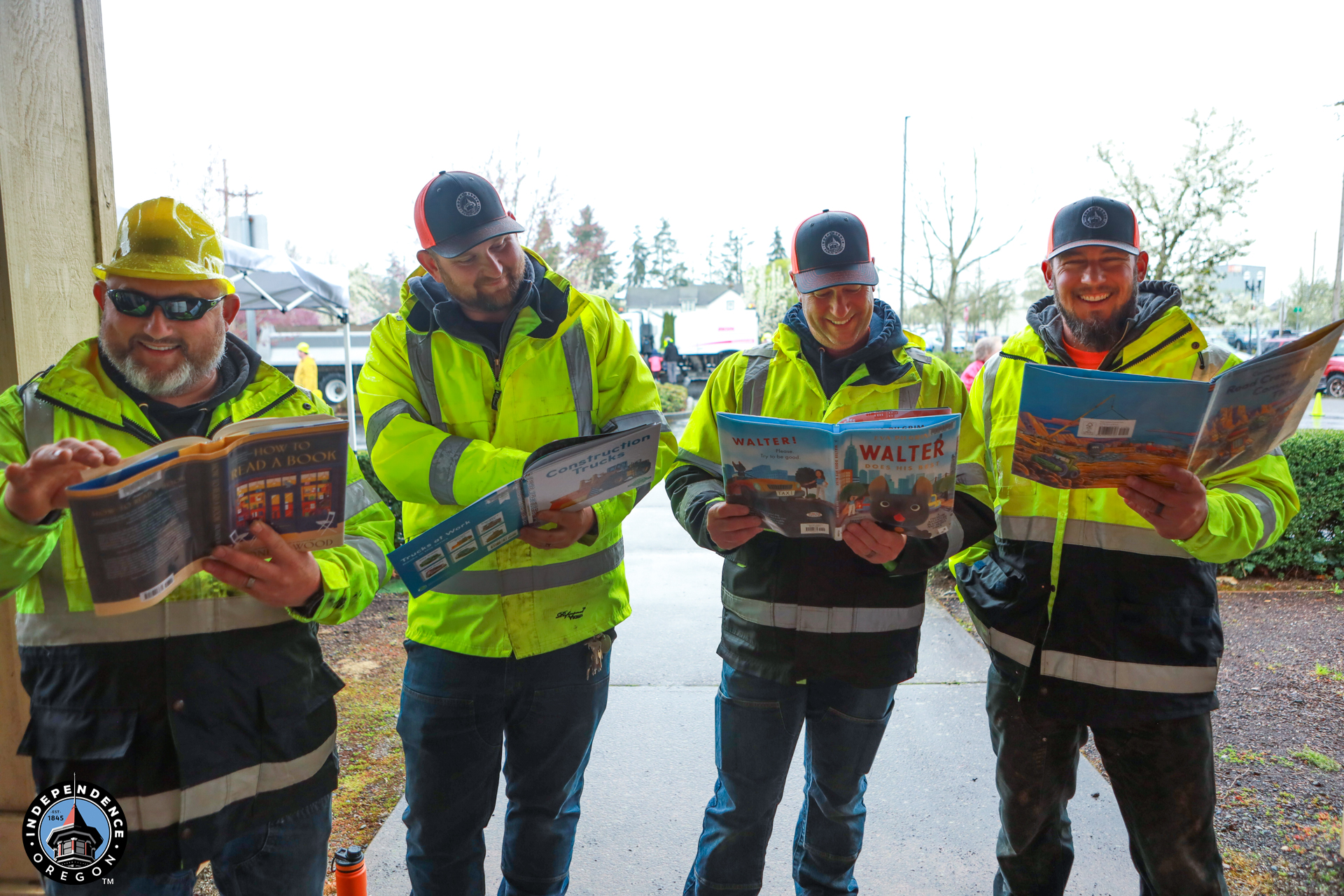 Four construction workers in bright yellow jackets and caps smile while reading children's books outdoors on a cloudy day, conveying joy and playfulness.