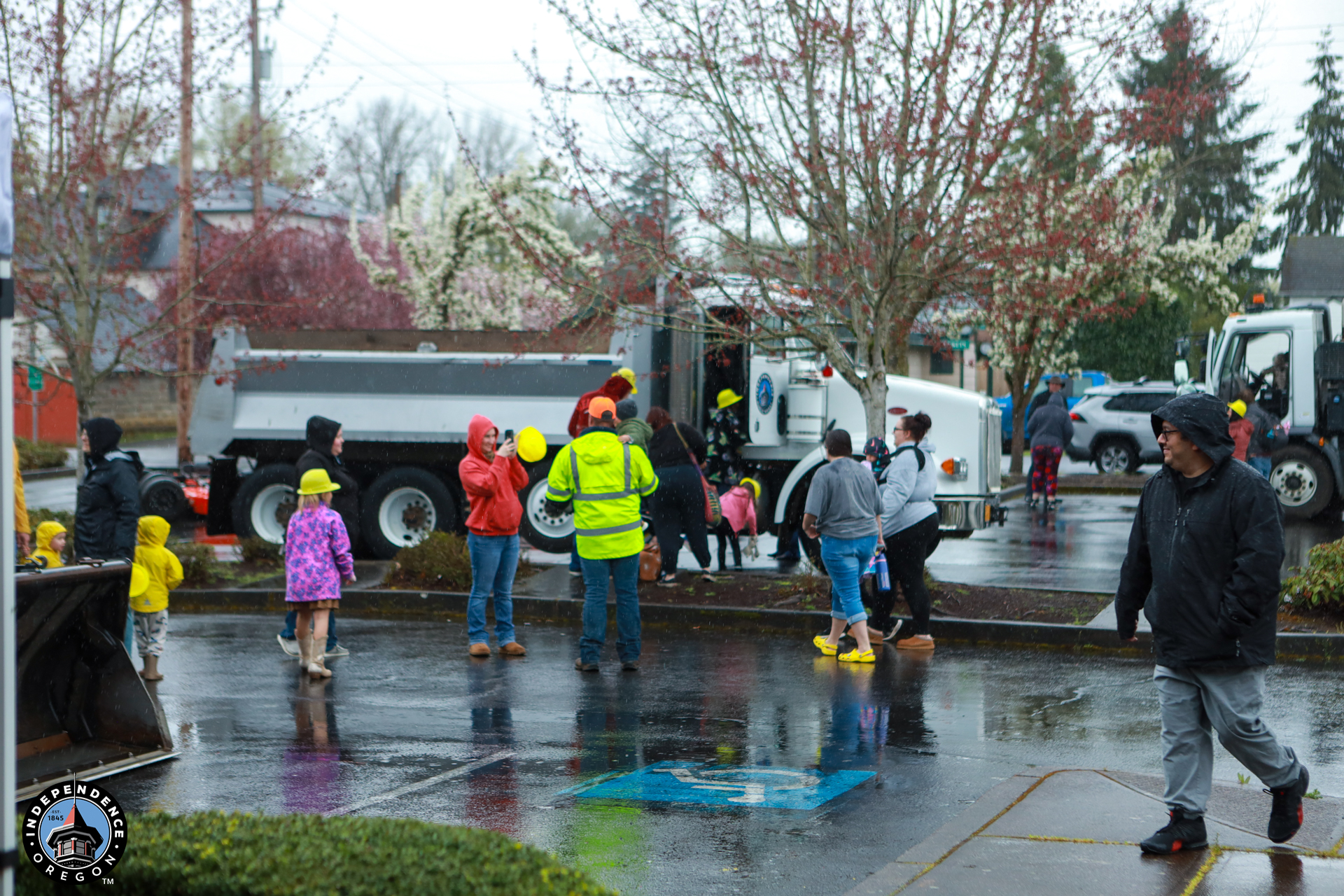 A rainy scene in a parking lot shows children and adults in colorful rain gear and helmets exploring utility trucks. The atmosphere is lively and curious.