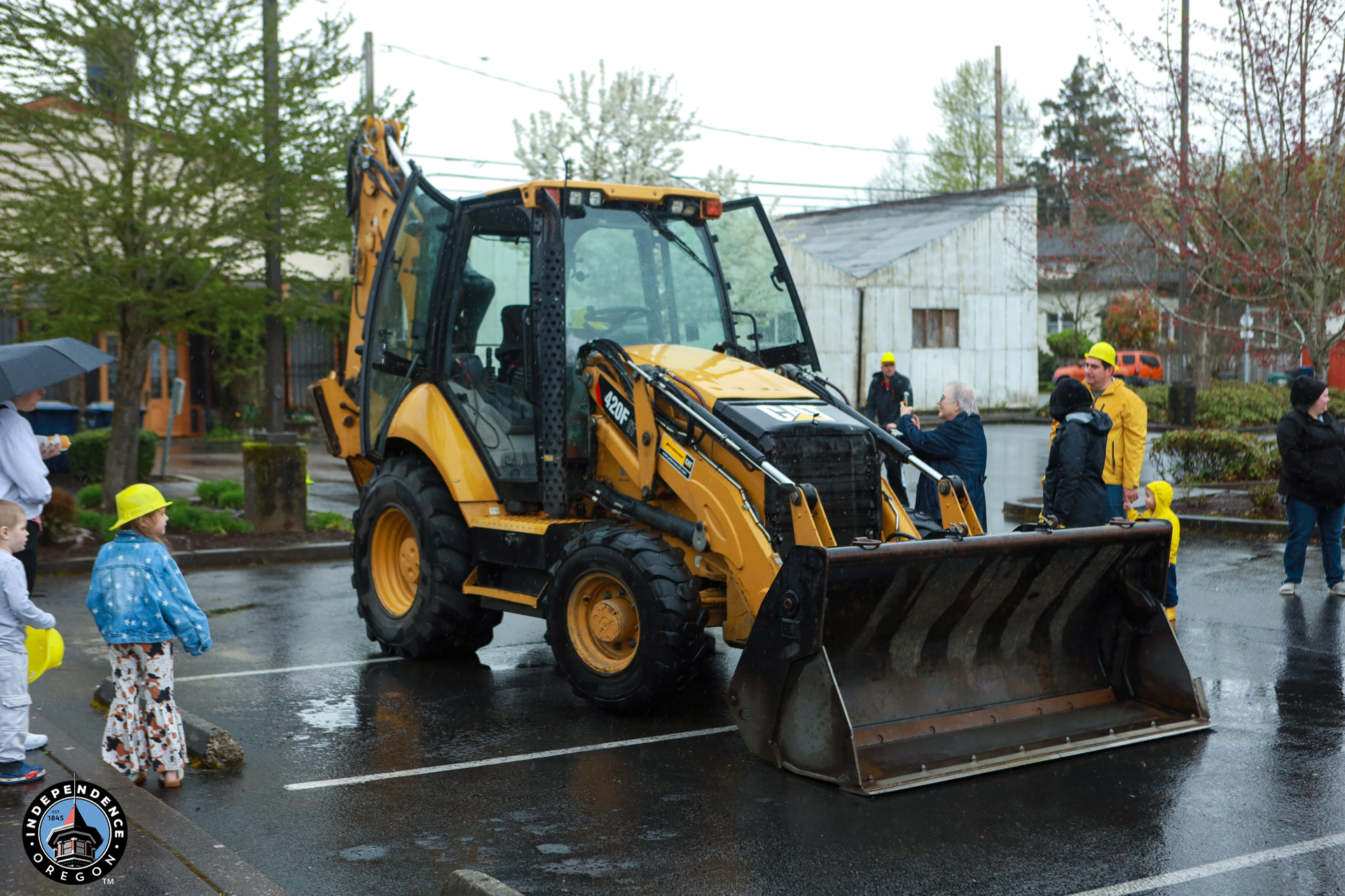 A yellow backhoe loader parked on wet pavement, surrounded by adults and children wearing raincoats and hats. Overcast skies suggest recent rain.