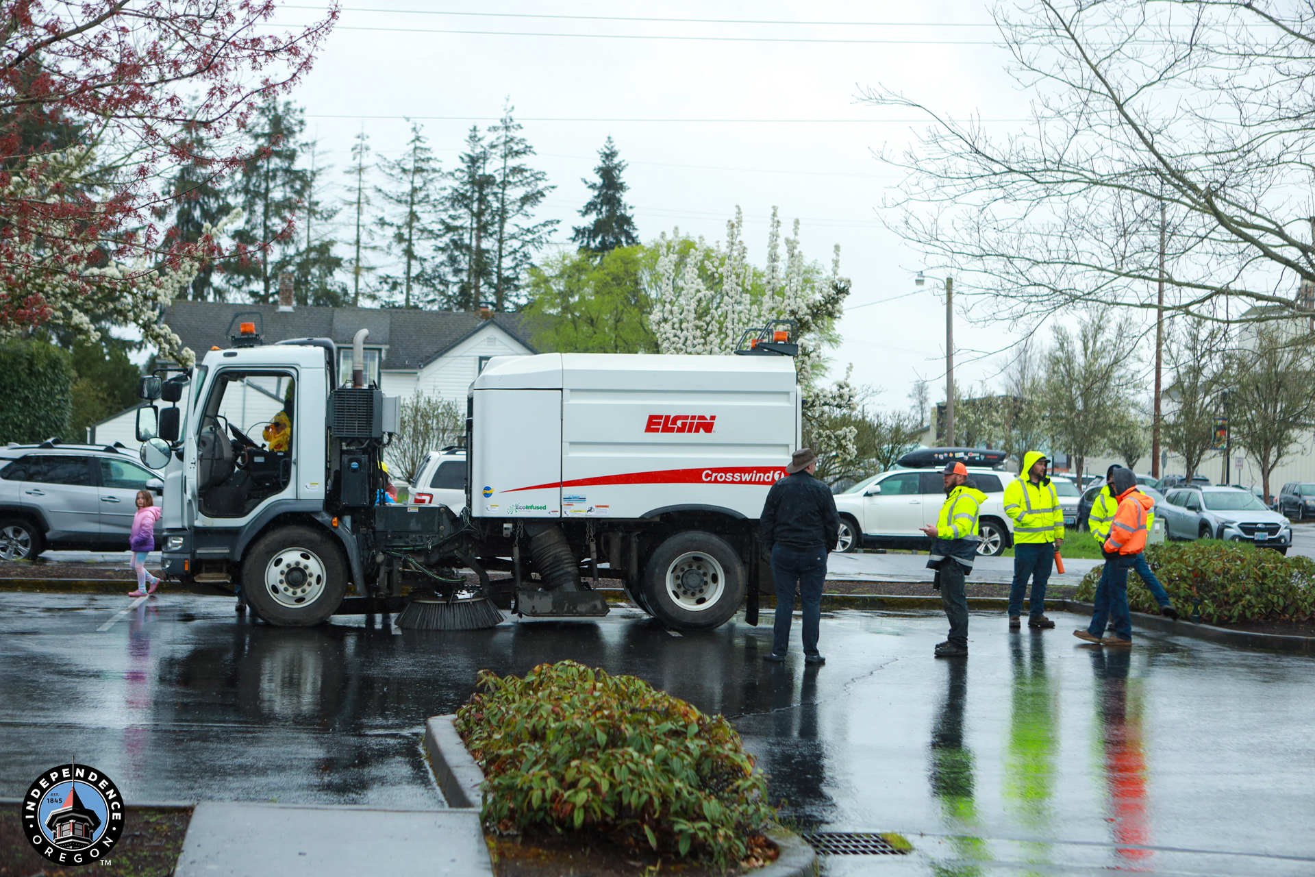 A street sweeper truck is parked on a wet road surrounded by four workers in high-visibility jackets. The setting is outdoors on a rainy day.