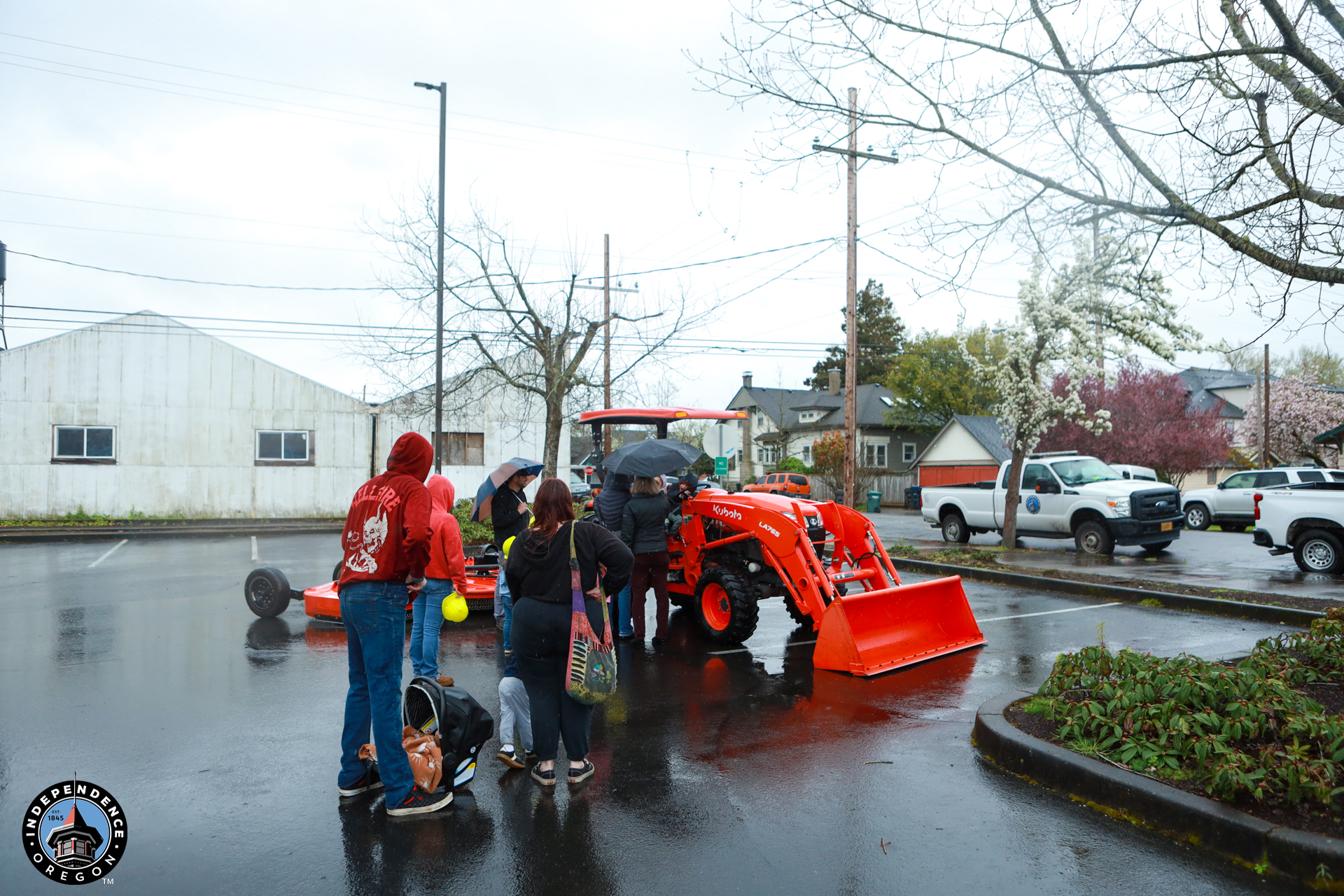 A group of people gather around an orange tractor in a rainy, suburban parking lot. Some hold umbrellas while children explore the scene. Wet pavement reflects the overcast sky.