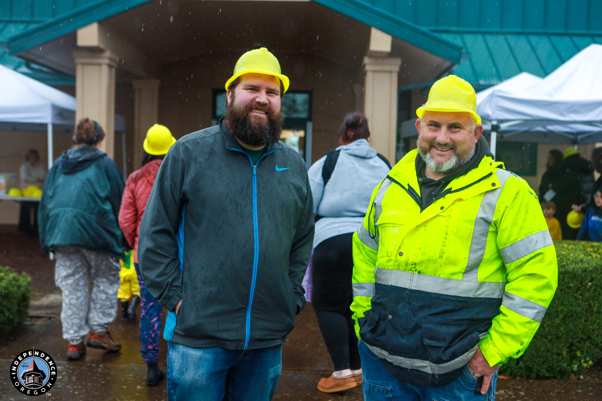 Two smiling men in yellow hard hats, one in a bright rain jacket, stand outside under a drizzly sky. Community event with tents and people in the background.