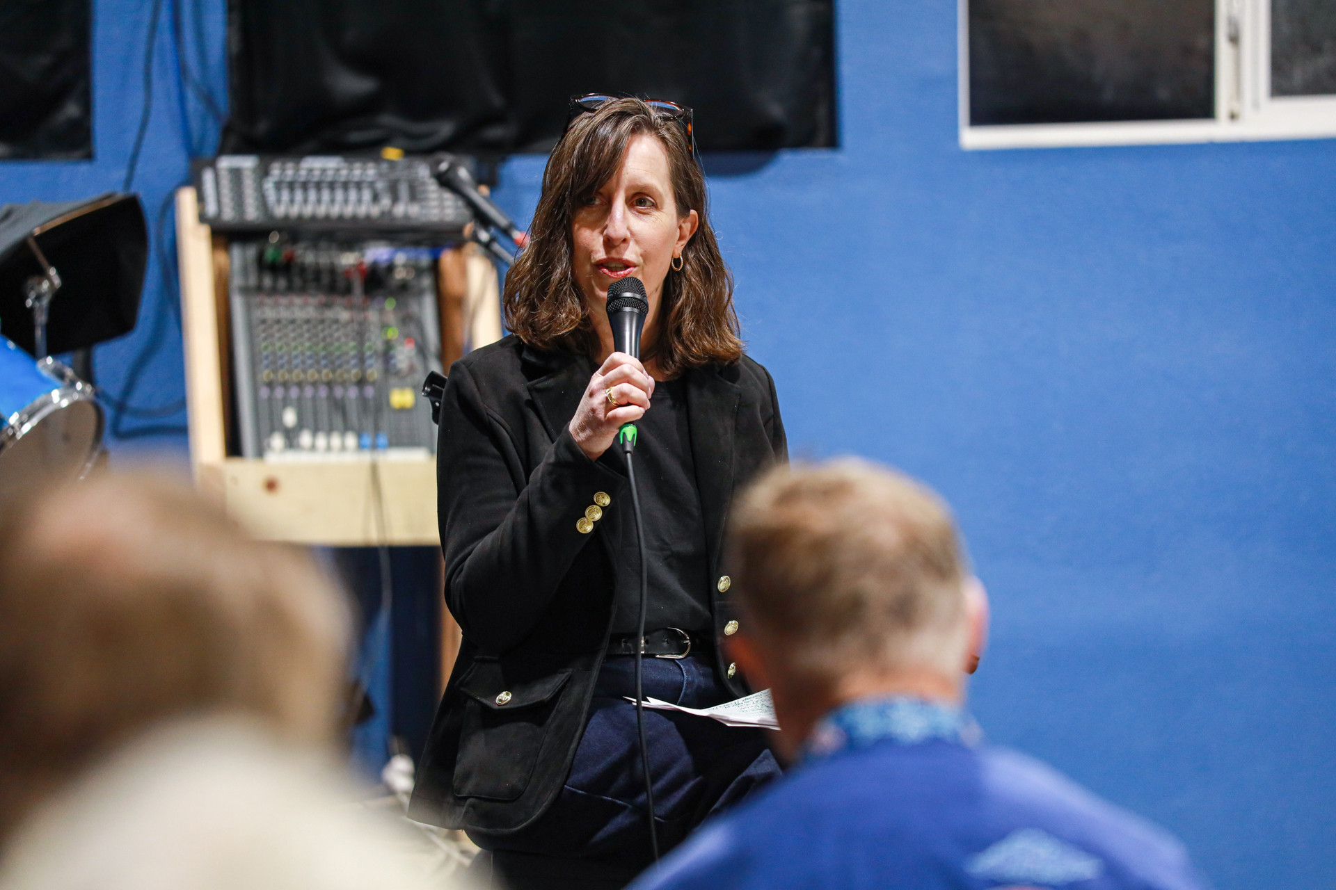 A woman in a black blazer speaks into a microphone, seated in front of a blue wall with sound equipment. Audience members with blurred heads listen intently.