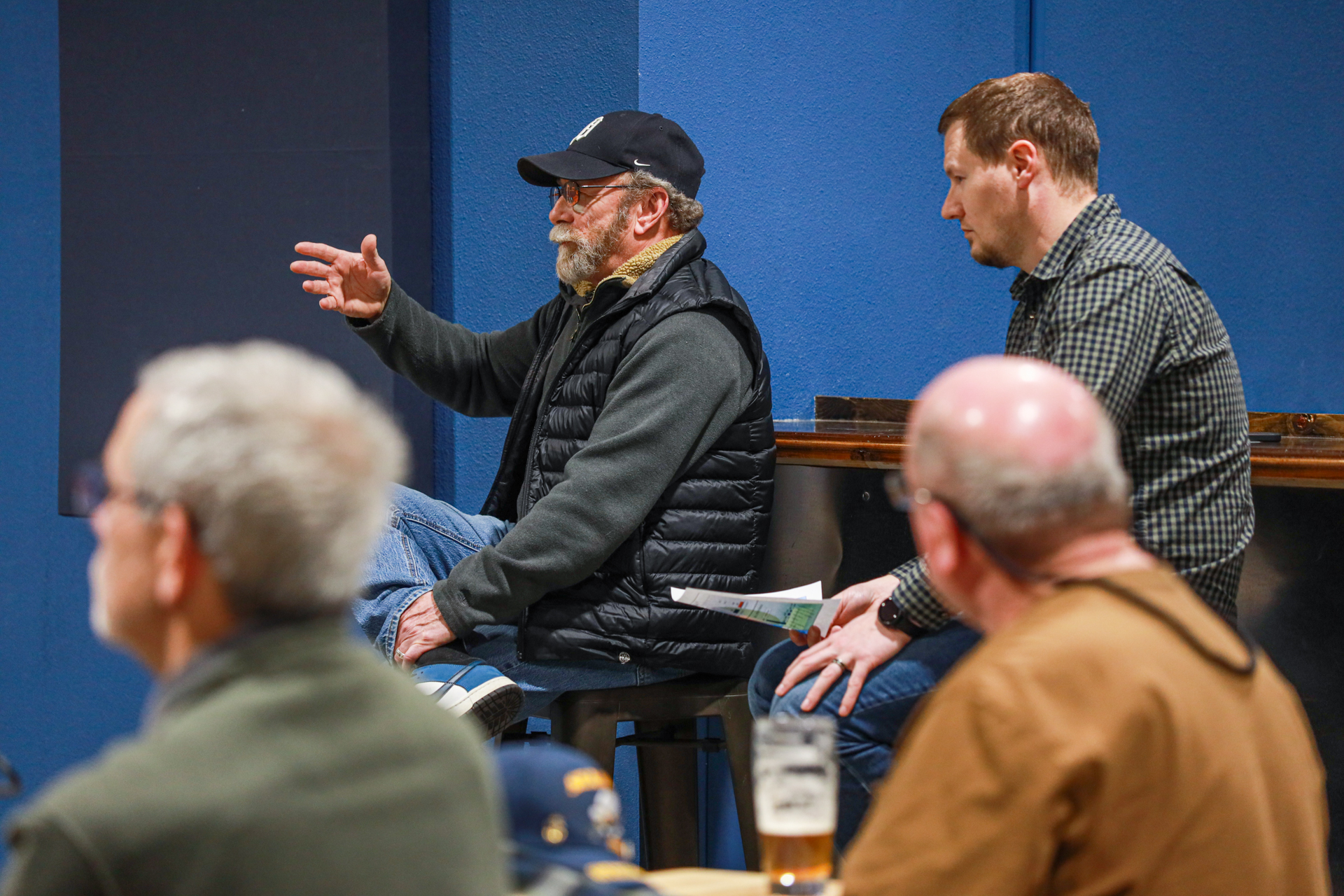 A man in a cap and vest gestures during a meeting in a blue-walled room. Others listen attentively, with drinks on the table, creating a casual, engaged atmosphere.