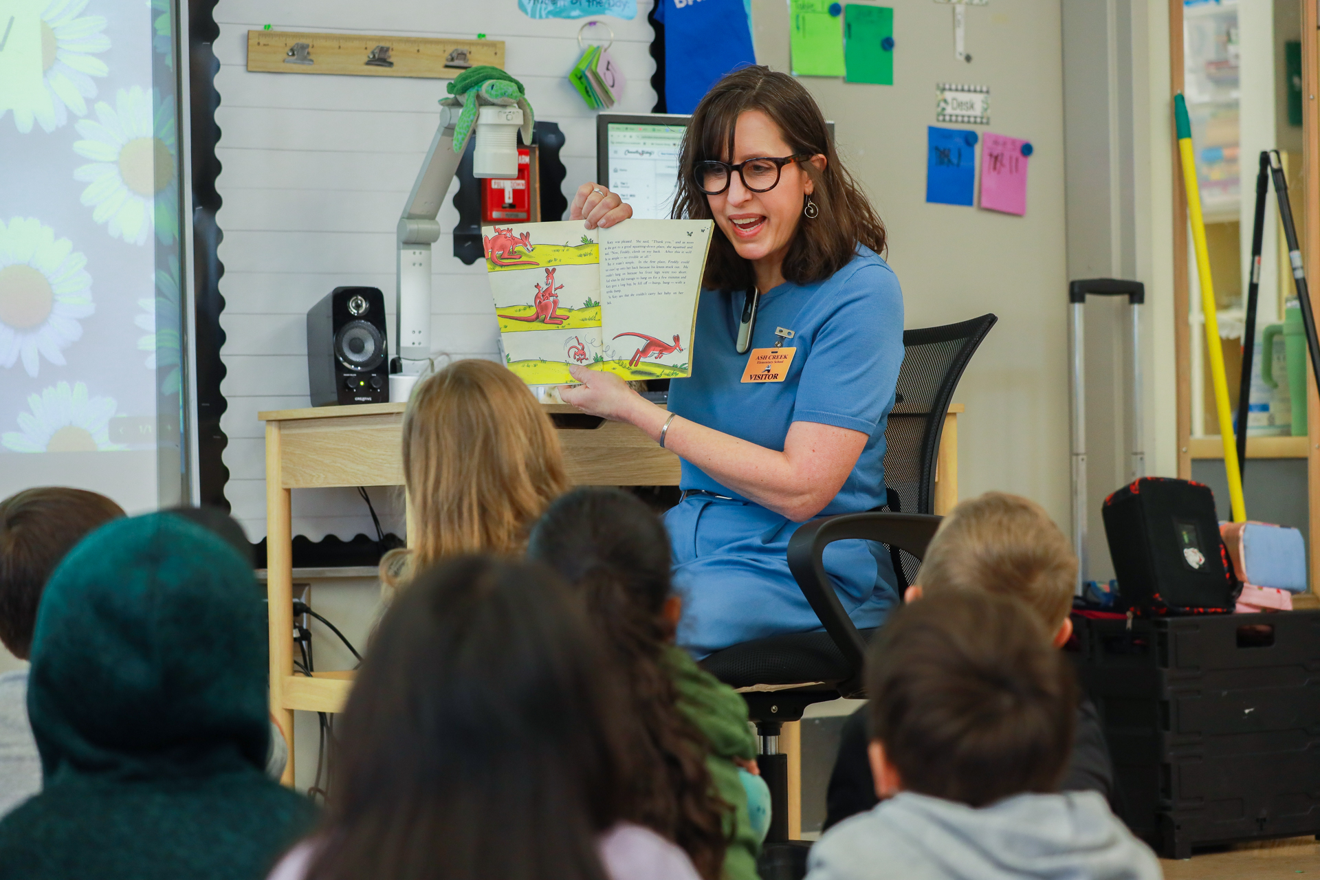 A woman in blue reads a colorful children's book to attentive young students in a classroom. The room is decorated with posters, conveying a vibrant atmosphere.