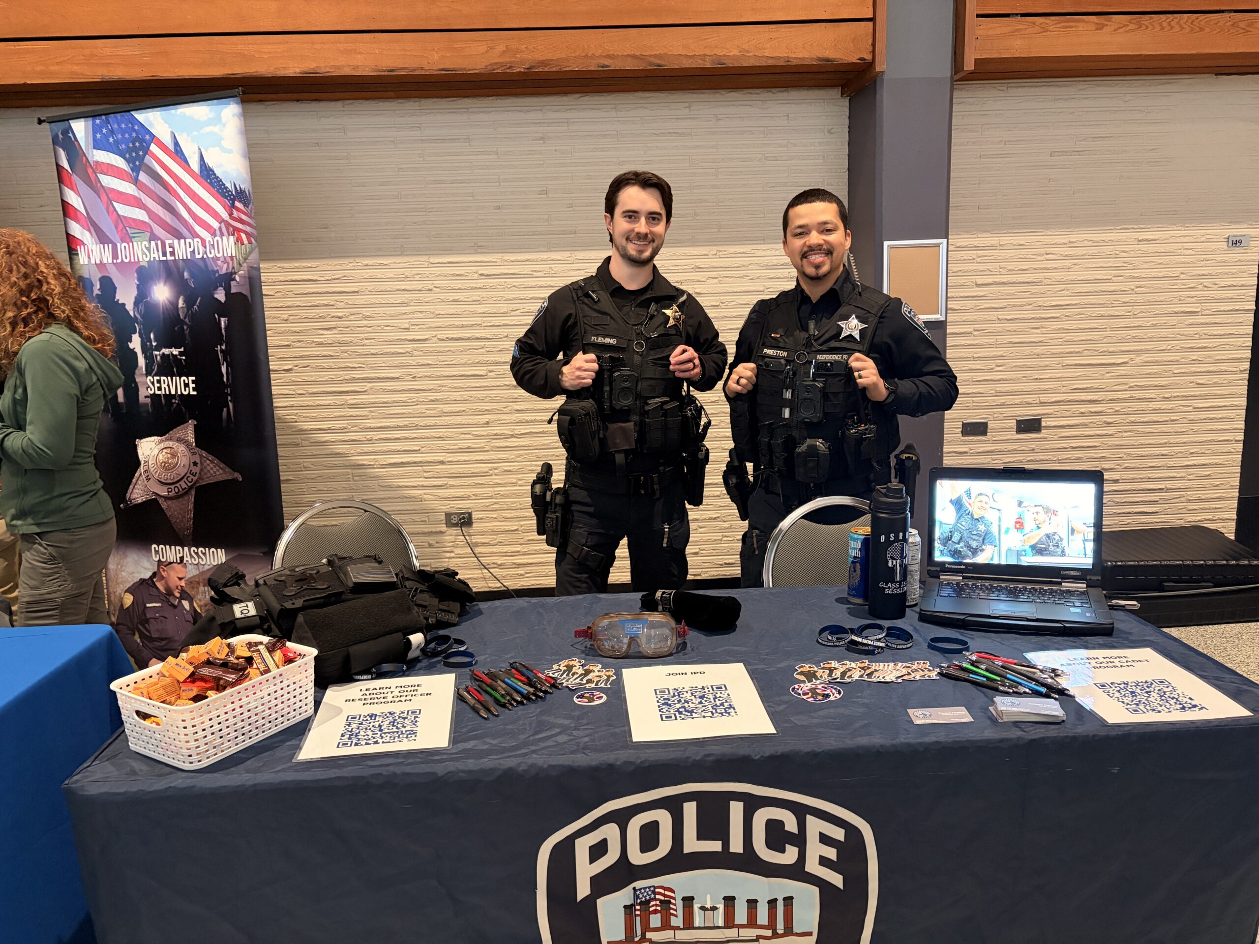 Two police officers stand smiling behind a recruiting booth. The table has QR codes, promotional items, and a laptop. A banner displays an American flag.