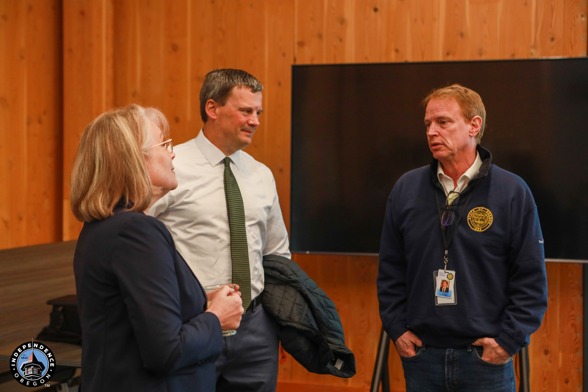 Three people stand talking in a wooden-paneled room. Two are in business attire, and one wears a navy jacket with a badge. The atmosphere is professional.