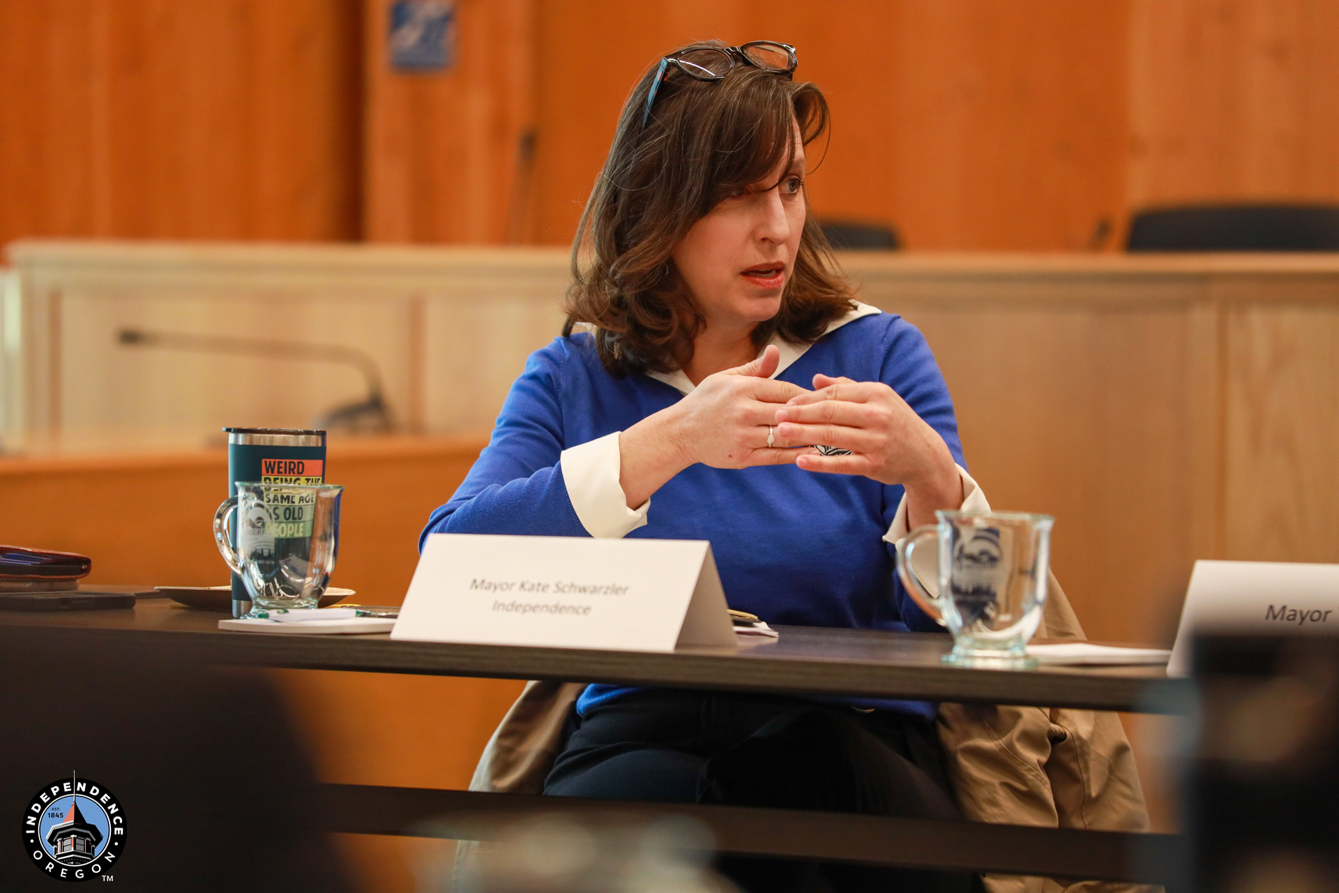 A woman in a blue top speaks animatedly at a conference table. A nameplate reads "Mayor Kate Schwarzer, Independence." Warm, engaged setting.