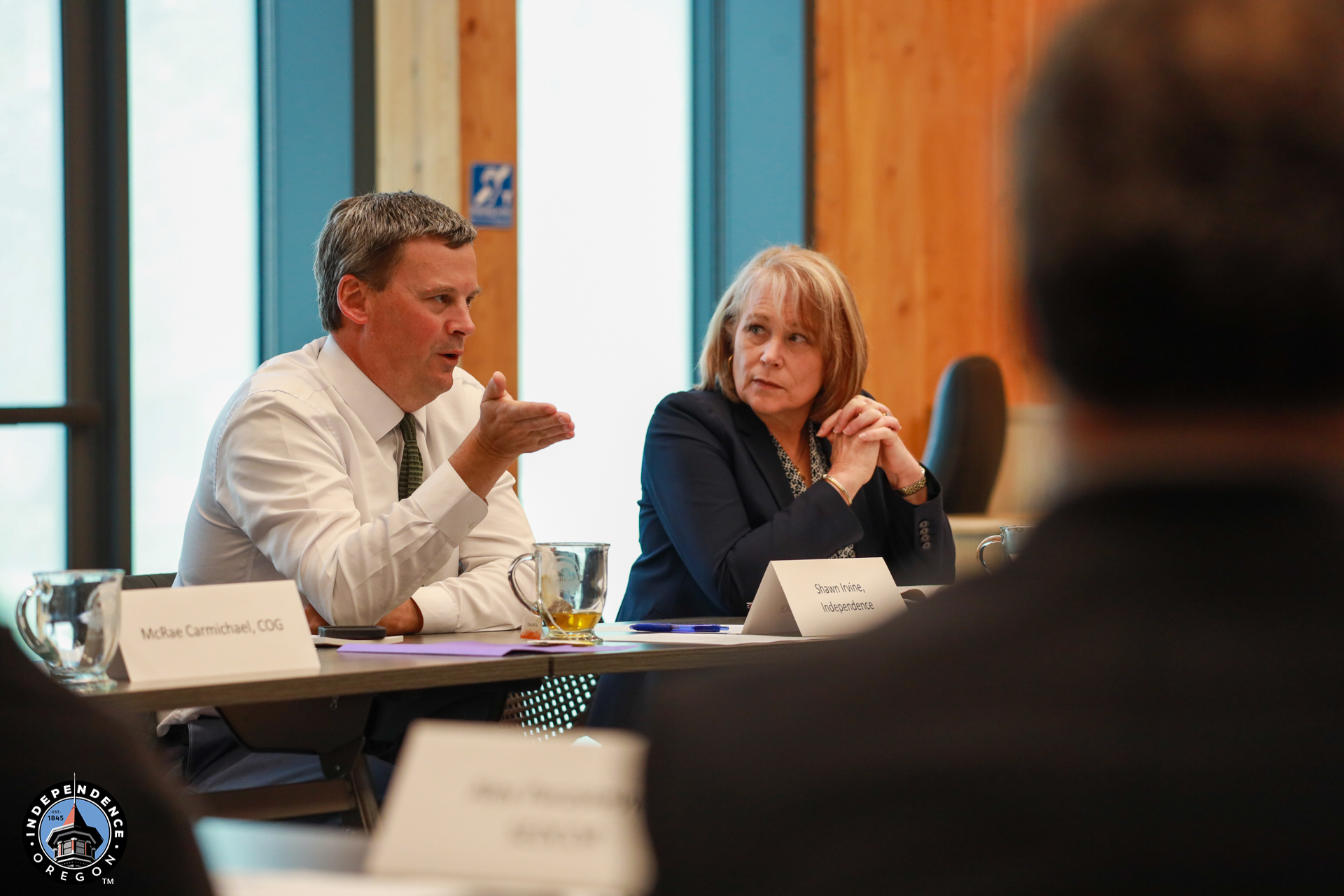 A man and a woman sit at a conference table, engaged in discussion. Papers and nameplates are visible. The mood is serious and focused.