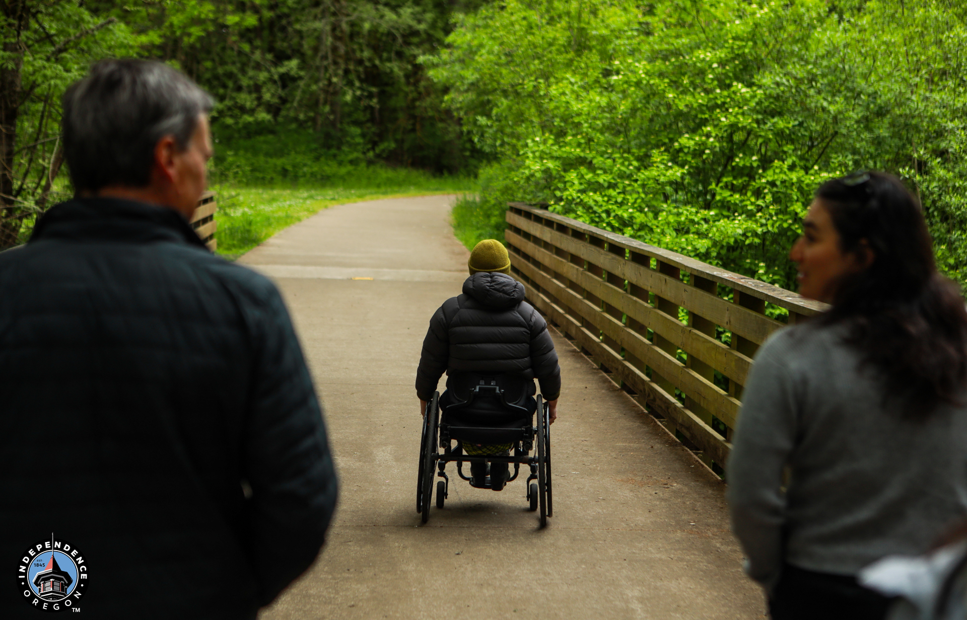 A person in a wheelchair travels on a paved path through a lush, green forest, flanked by two others walking beside them. The mood is peaceful and inclusive.