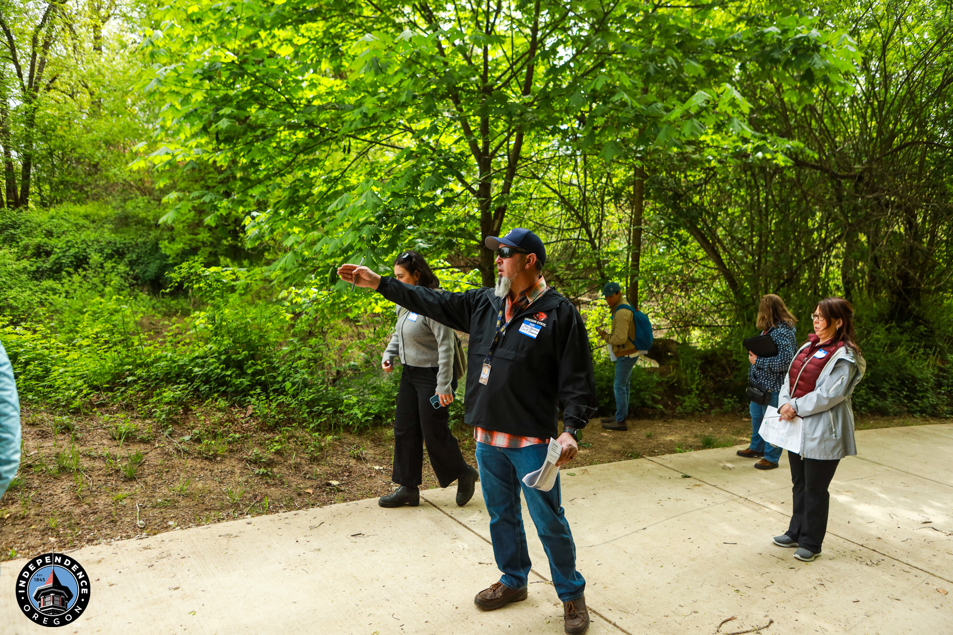 A group of people stands on a path in a lush, green forest. A man gestures, appearing to lead or guide. The scene feels educational and peaceful.