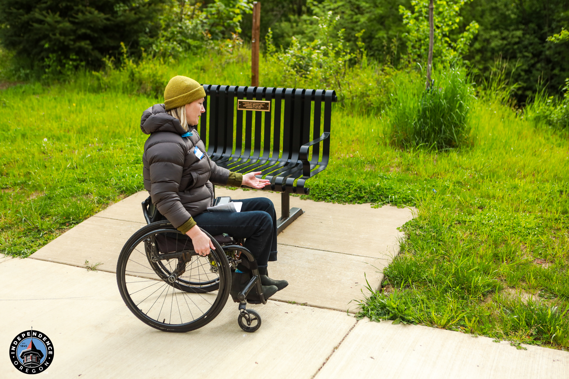 A person in a wheelchair on a concrete path gestures toward an empty bench surrounded by lush greenery, wearing a beanie and winter coat.