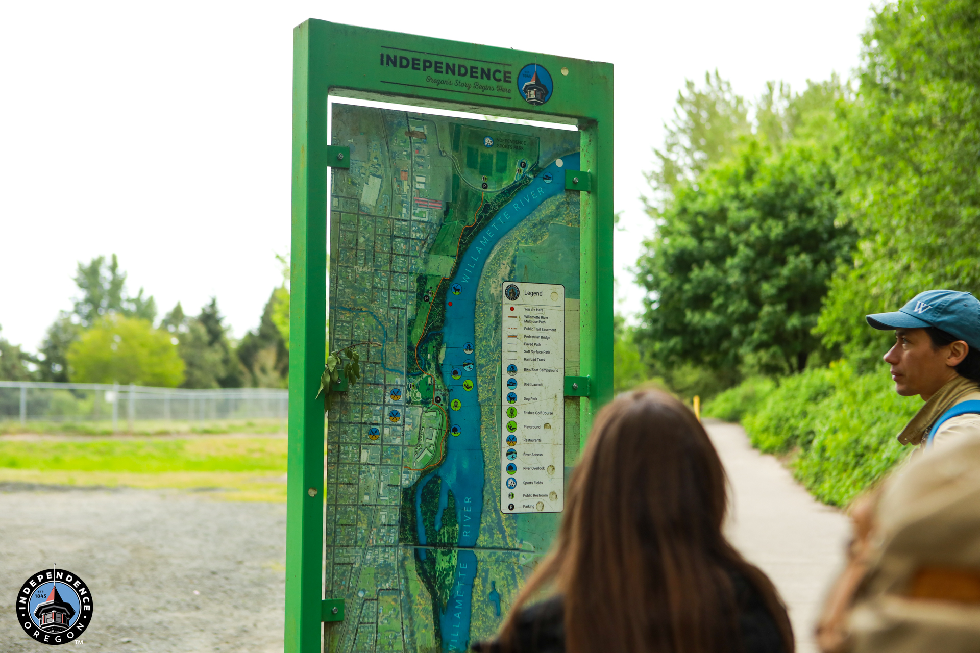 A group of people looks at a large, green-framed outdoor map labeled "Independence," showing a river and surrounding area. Lush trees line the pathway.