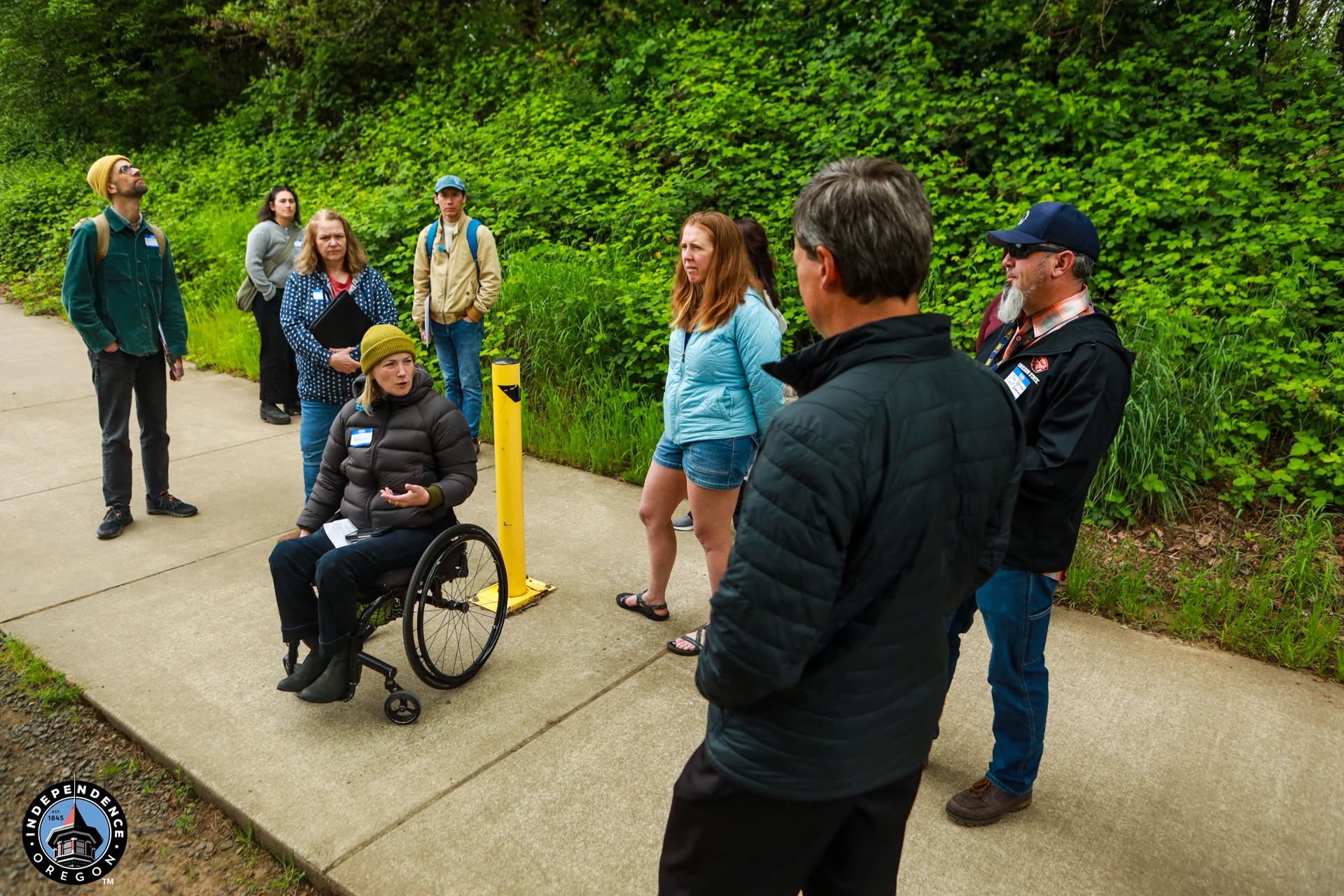 A group is gathered outdoors on a path lined with lush green foliage. A person in a wheelchair speaks to the group, creating an engaged and attentive atmosphere.