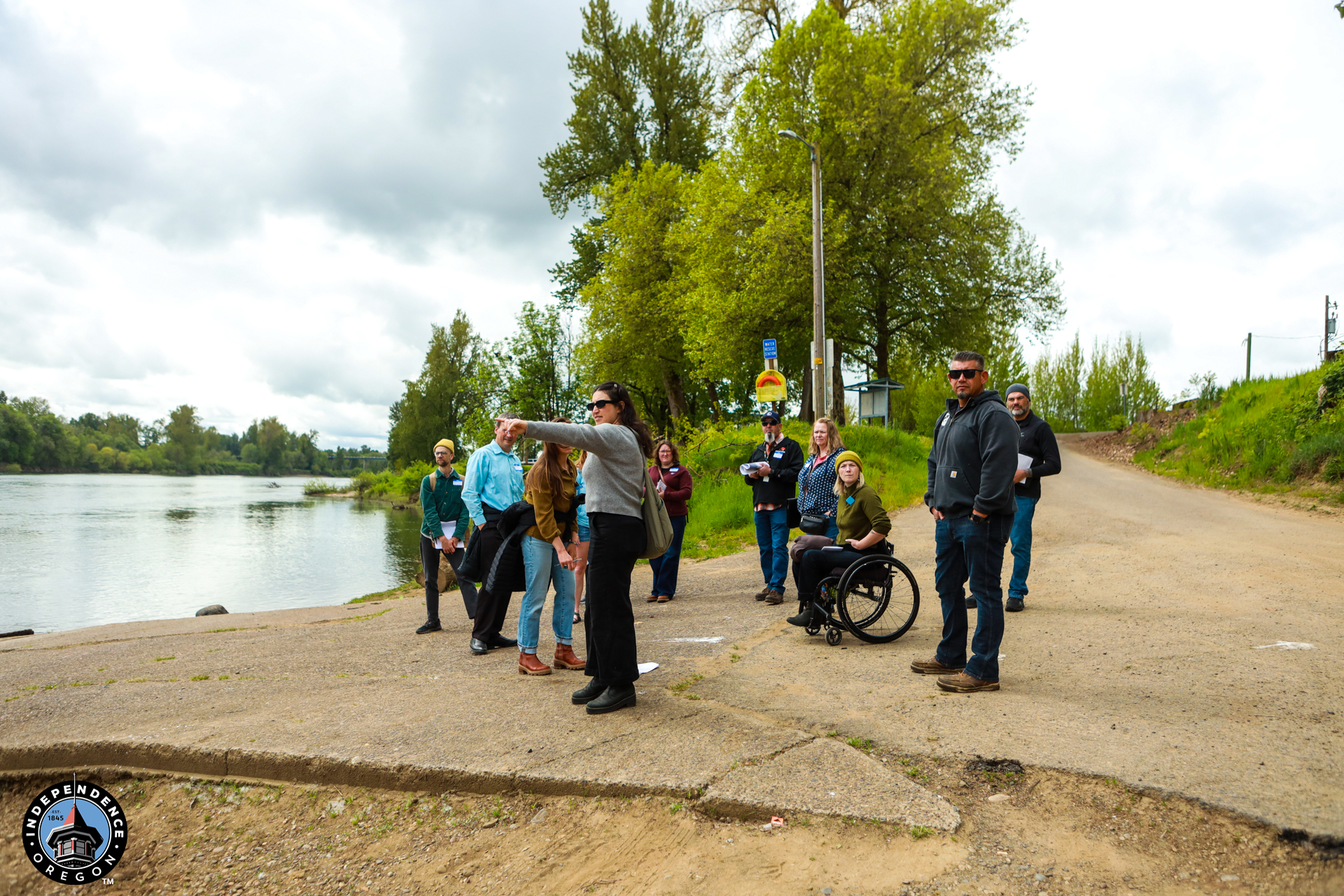 A group of people gather by a river under cloudy skies, some pointing or observing. Trees and a path are visible. The mood is peaceful and engaged.