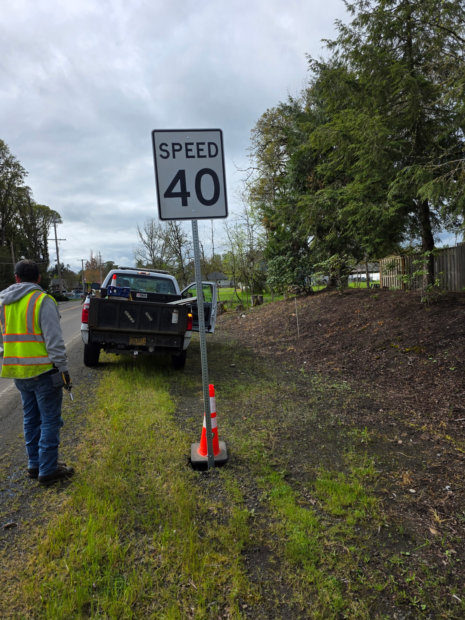 A person in a safety vest installs a "Speed 40" sign beside a rural road. A pickup truck is parked nearby under an overcast sky, with tall trees in the background.