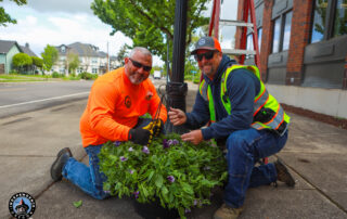 Two workers in bright safety gear kneel on a sidewalk, smiling and holding a hanging flower basket. A ladder and streetlamp are in the background.