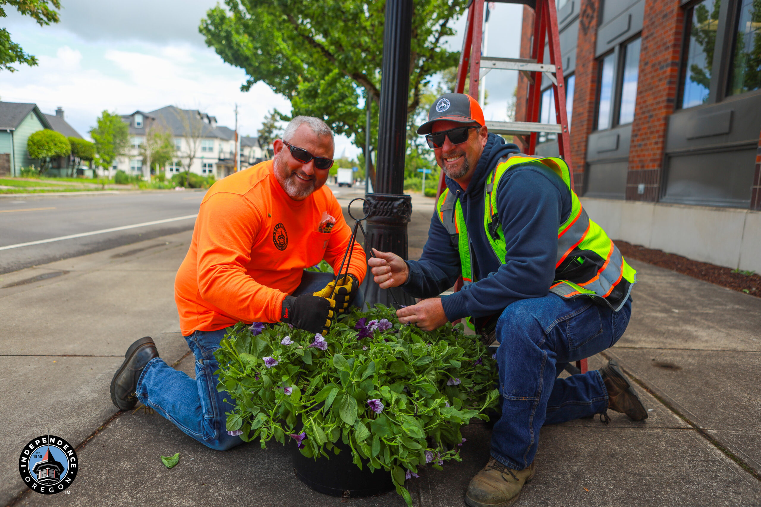 Two workers in bright safety gear kneel on a sidewalk, smiling and holding a hanging flower basket. A ladder and streetlamp are in the background.