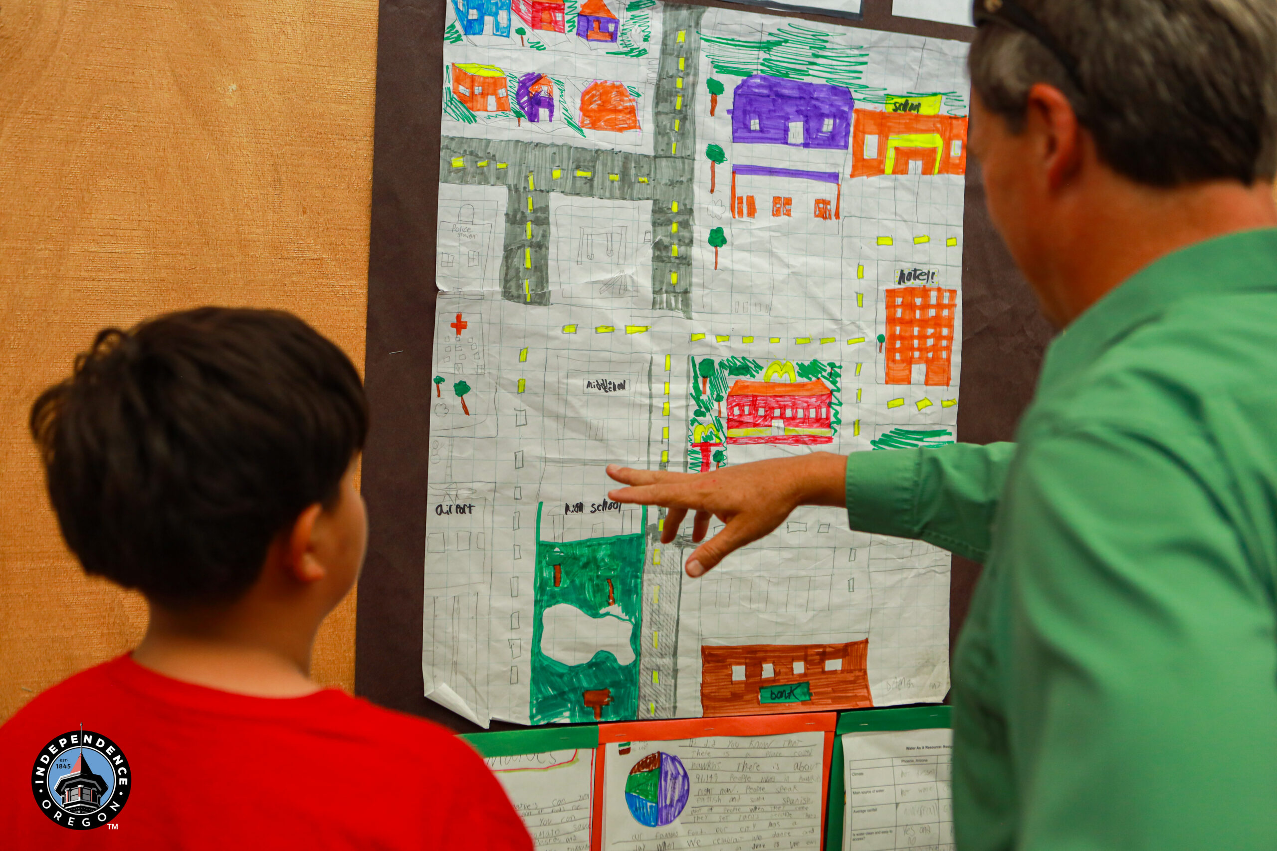 A child and an adult discussing colorful artwork on a wall, showcasing creative projects and maps in a classroom setting.