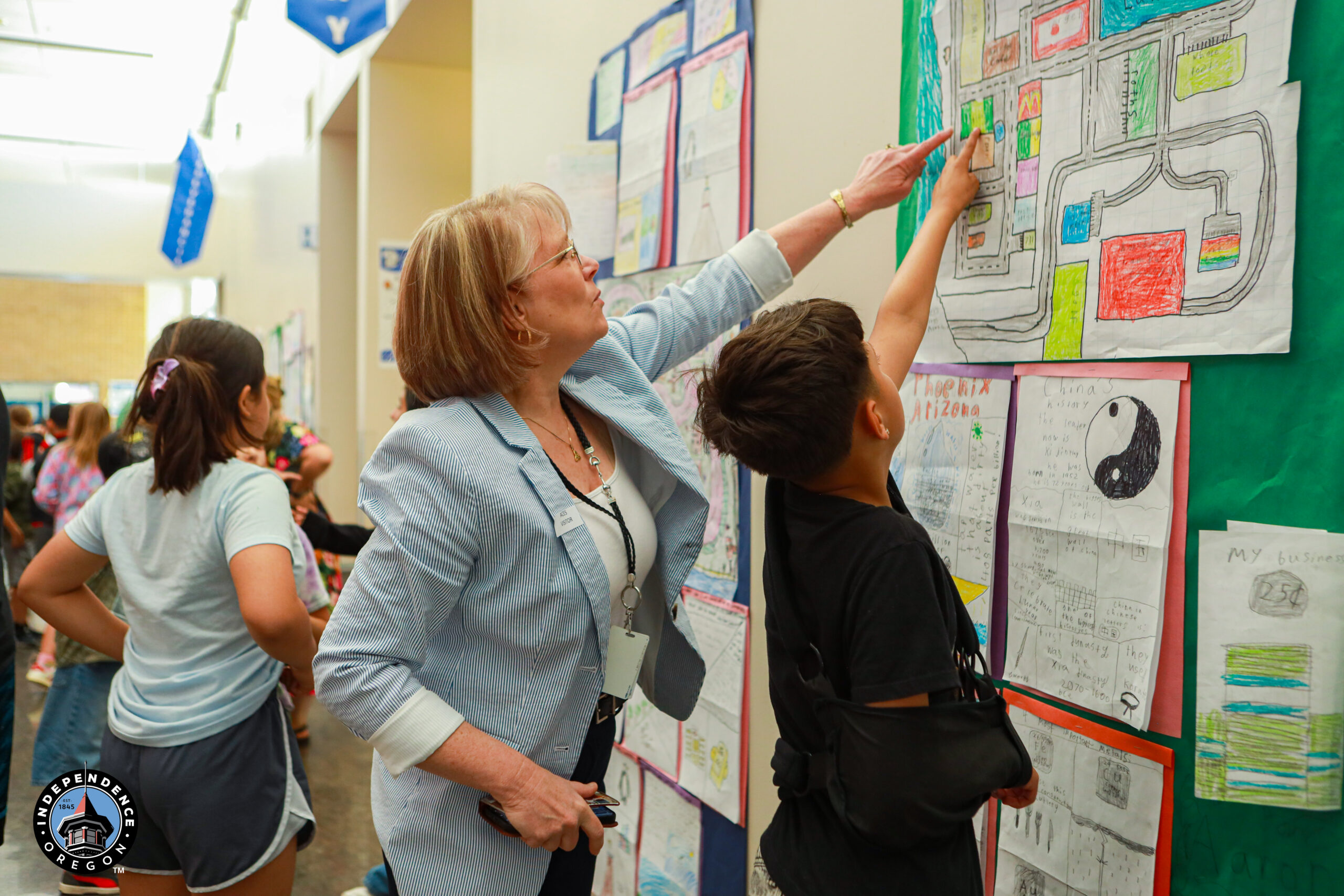 A woman engages with a student, pointing at colorful drawings on a classroom wall during an educational event.