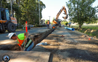 Construction workers in safety gear dig a long trench on a sunlit street, with a backhoe nearby and blue pipes ready for installation, amid lush trees.