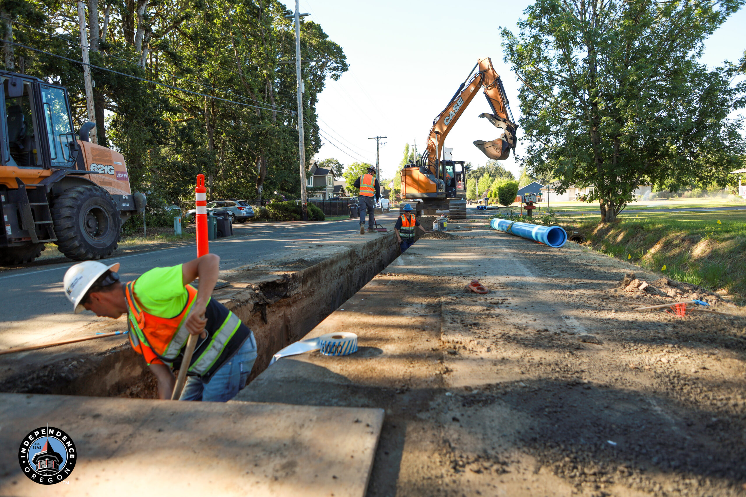 Construction workers in safety gear dig a long trench on a sunlit street, with a backhoe nearby and blue pipes ready for installation, amid lush trees.