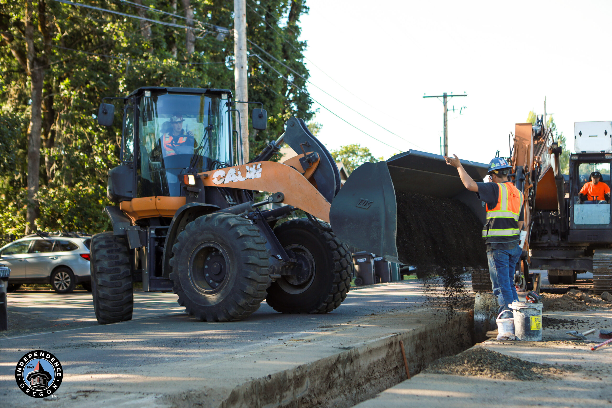 A construction worker in a safety vest gestures to an excavator pouring gravel into a trench on a sunlit street, with trees and utility poles nearby.