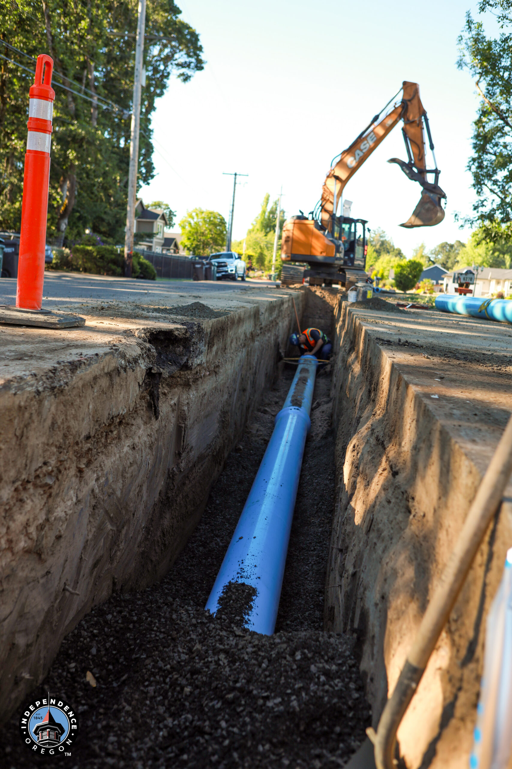 A large excavator installs blue piping in a deep trench along a street, with a worker nearby. Bright cones and trees line the road, suggesting active construction.