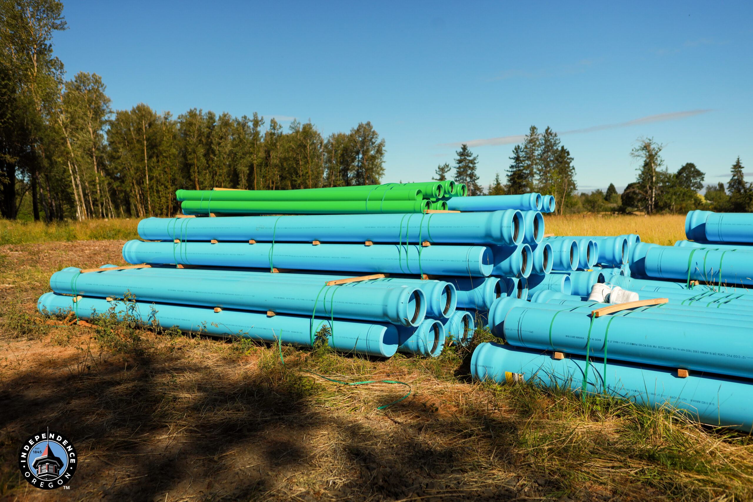 Stacks of large blue and green pipes lie on dry grass in a field, bordered by trees under a clear blue sky, creating a calm, organized scene.