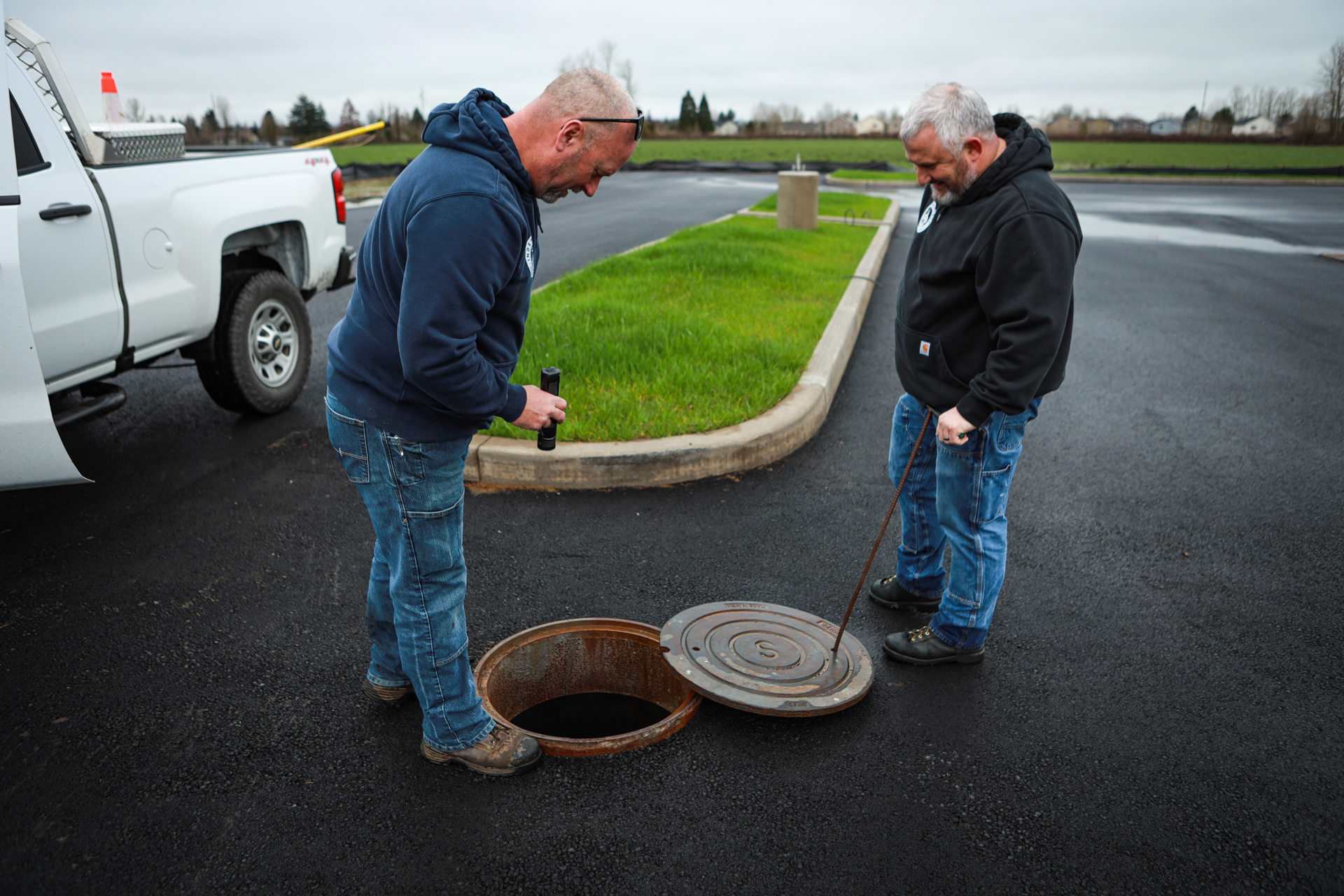 Two men inspecting an open manhole on a paved road near a white pickup truck and grassy curb.
