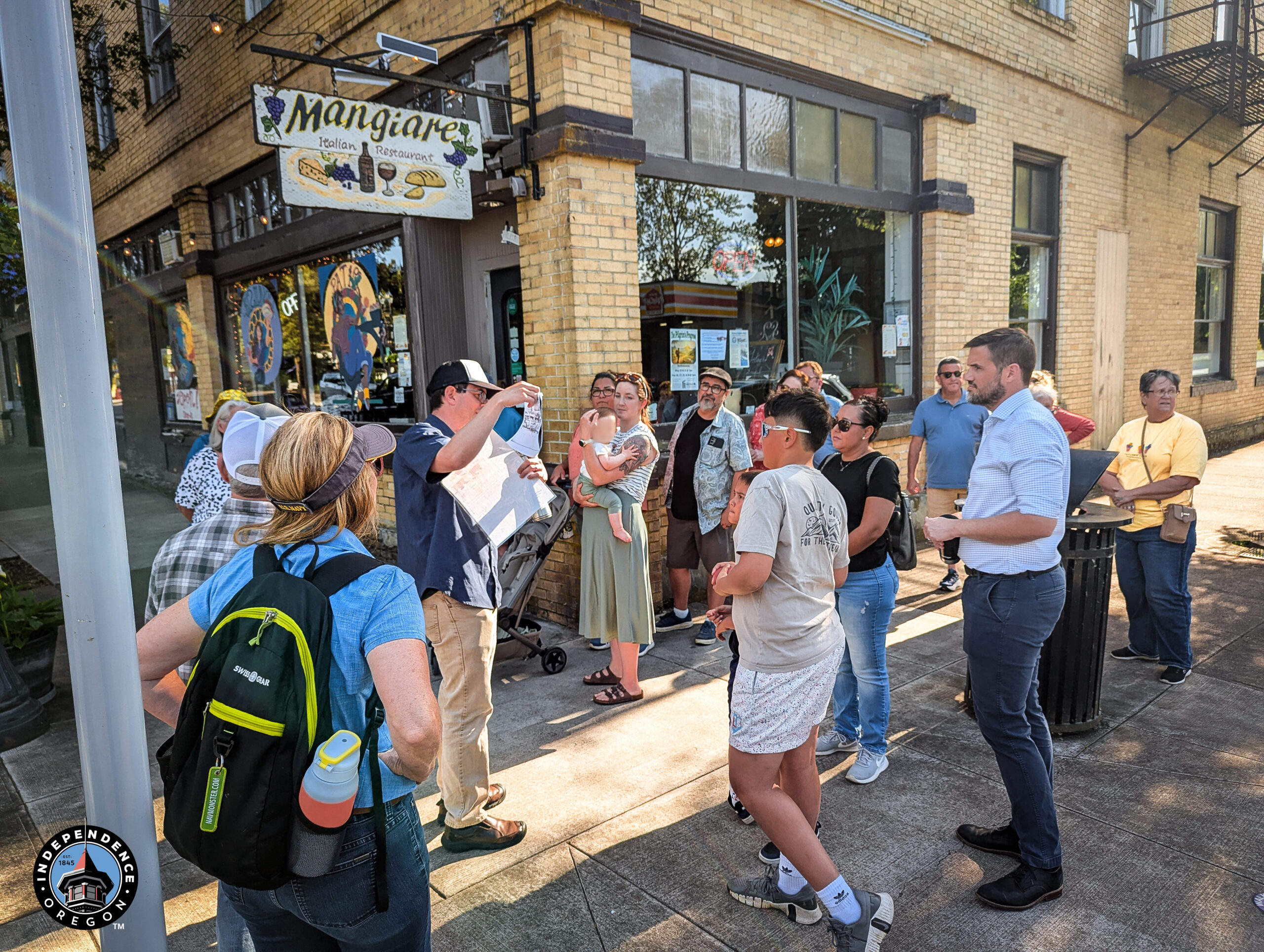 A group of people stands outside a brick building with a "Mangiare Italian Restaurant" sign. A person in a cap gestures while holding a document. The mood is engaged and attentive.