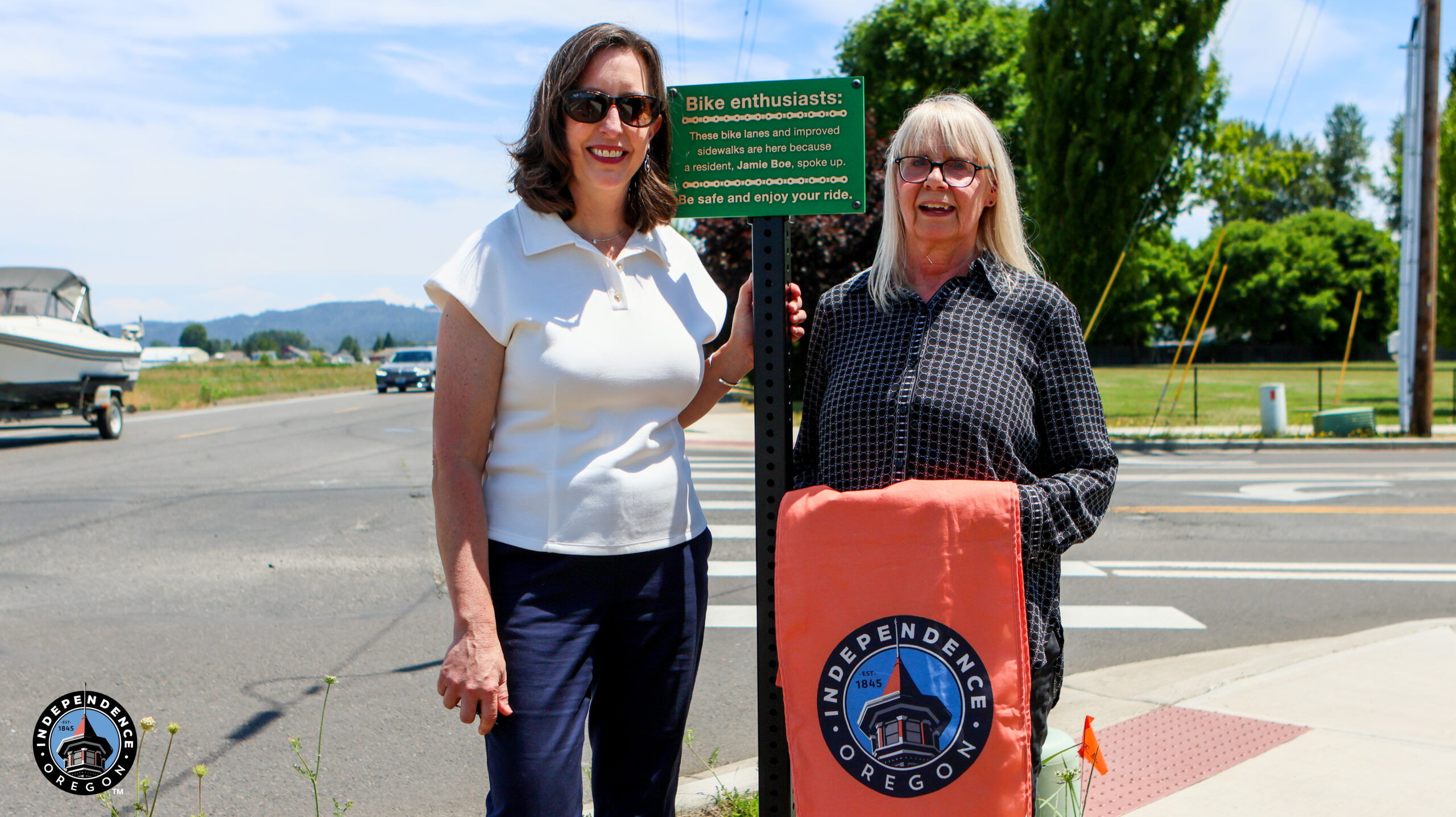 Two women stand beside a bike safety sign in Independence, Oregon, with a boat in the background and trees lining the street.