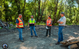 Four city workers in safety vests discussing on a cleared site surrounded by trees and debris.