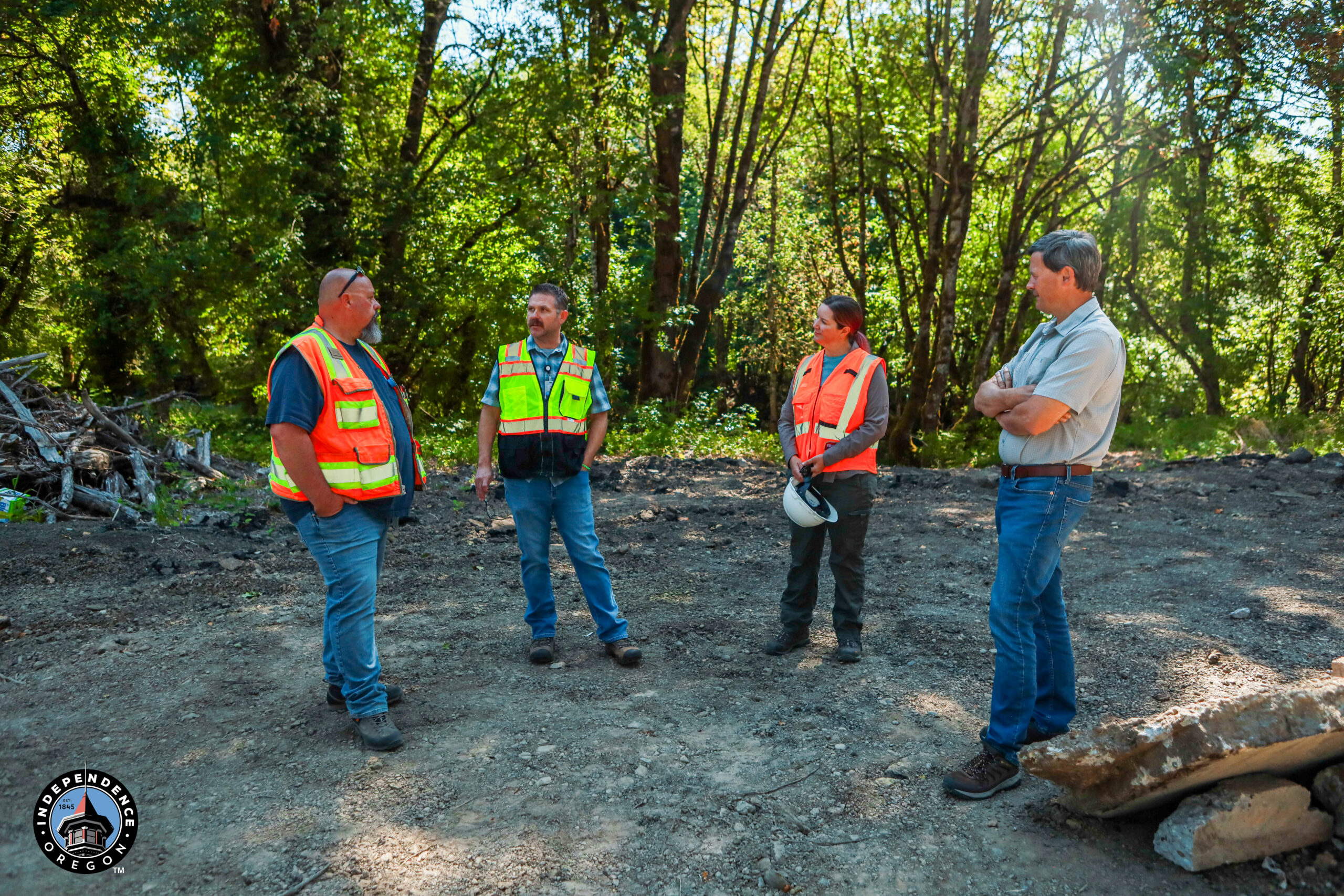 Four city workers in safety vests discussing on a cleared site surrounded by trees and debris.