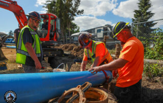 Three construction workers in high-visibility vests and hard hats work on a large blue pipe in a dirt trench near an orange Kubota excavator.