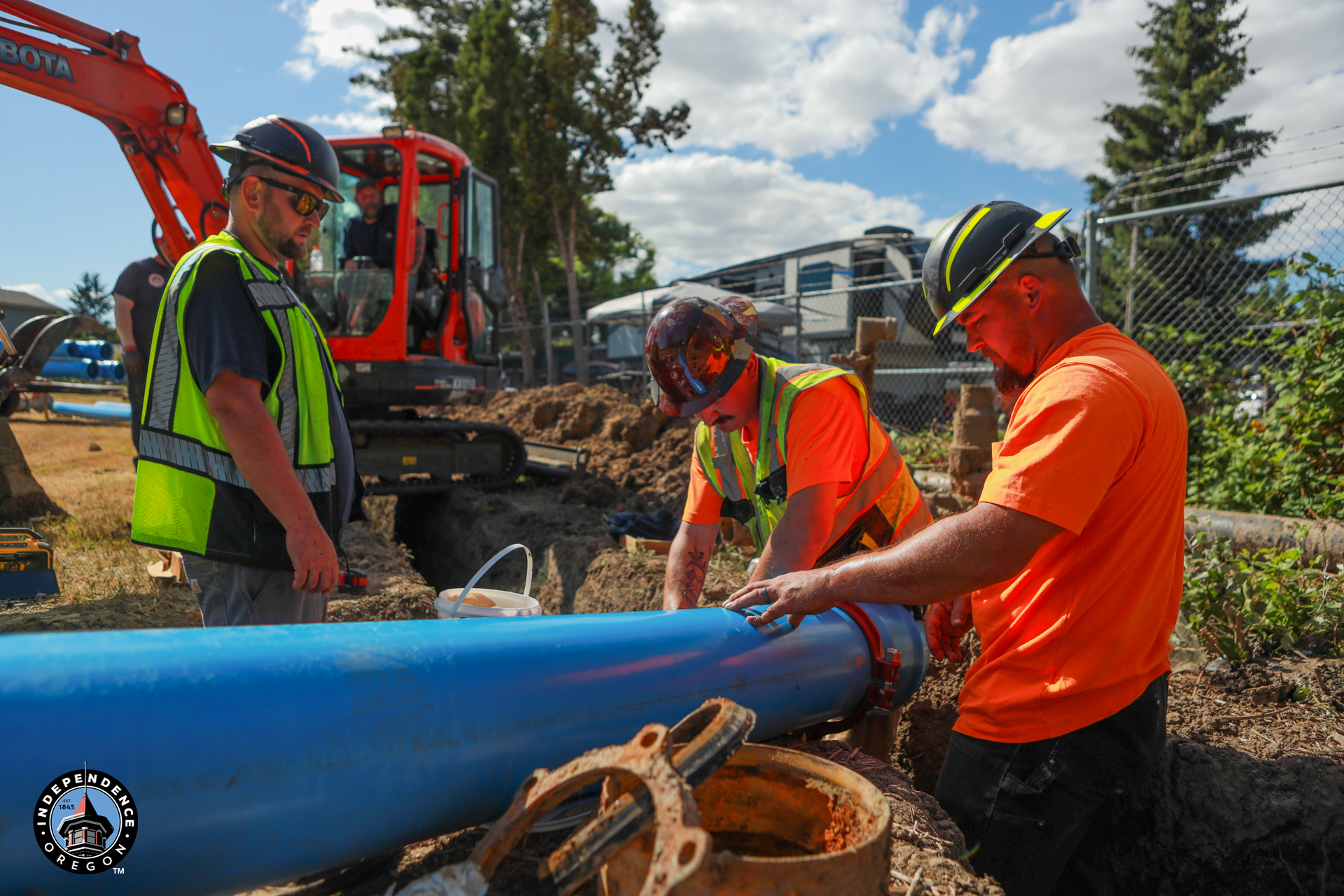 Three construction workers in high-visibility vests and hard hats work on a large blue pipe in a dirt trench near an orange Kubota excavator.
