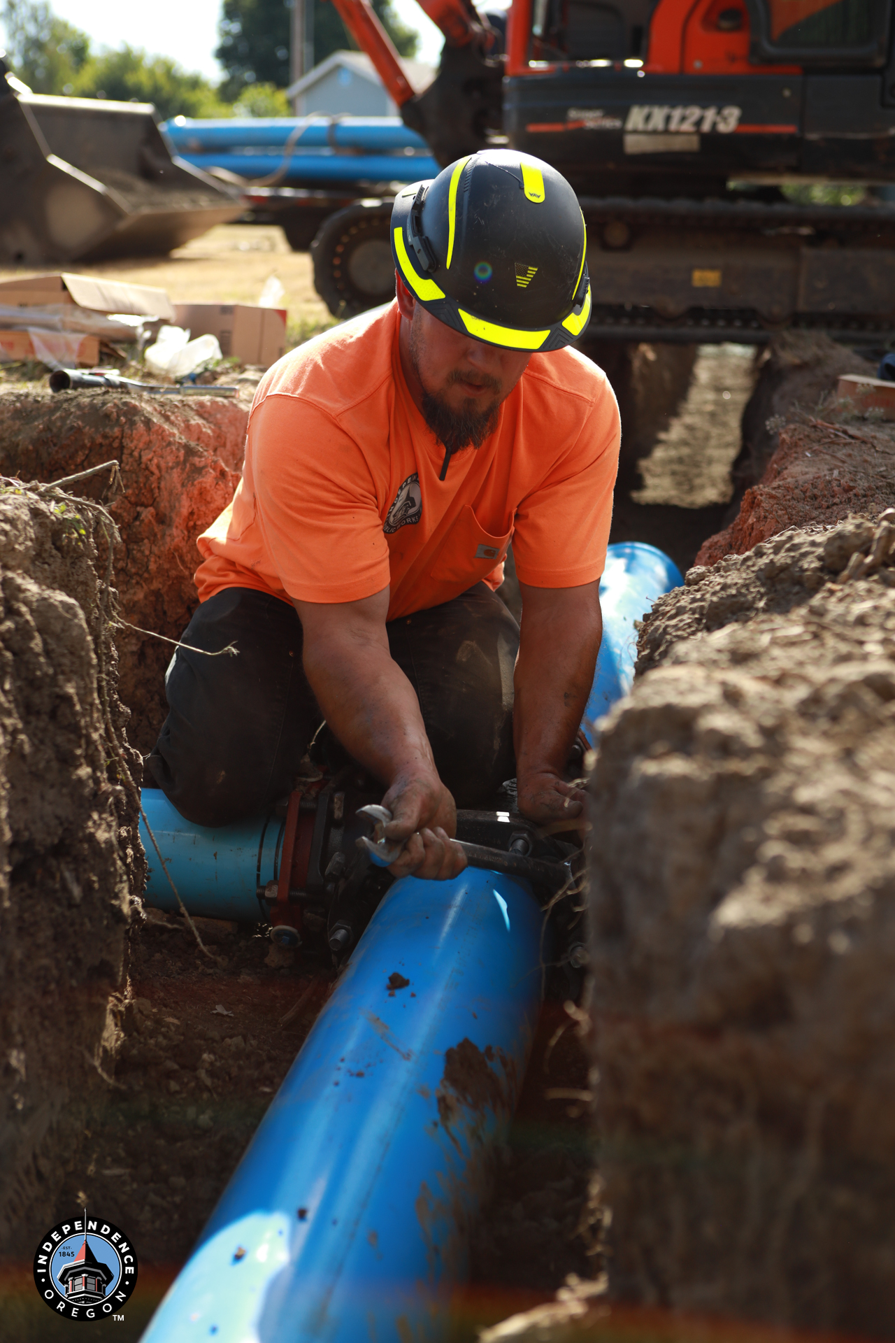 A construction worker in an orange shirt and safety helmet kneels in a dirt trench, using a wrench to tighten a bolt on a large blue water pipe.