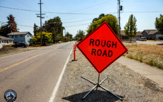 Bright orange “ROUGH ROAD” warning sign stands beside a dusty residential street, with utility poles, homes, trees, and cones visible.