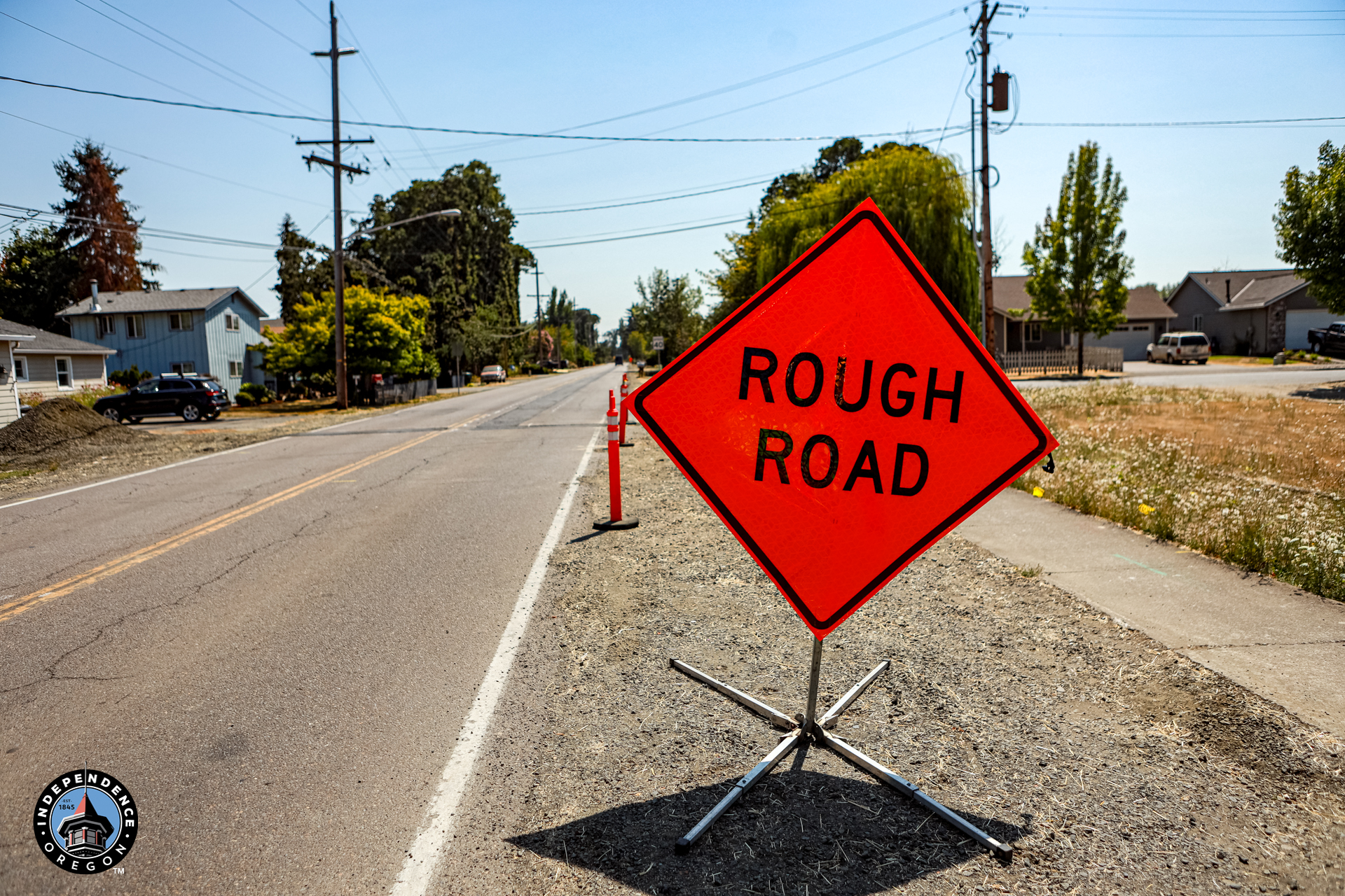 Bright orange “ROUGH ROAD” warning sign stands beside a dusty residential street, with utility poles, homes, trees, and cones visible.